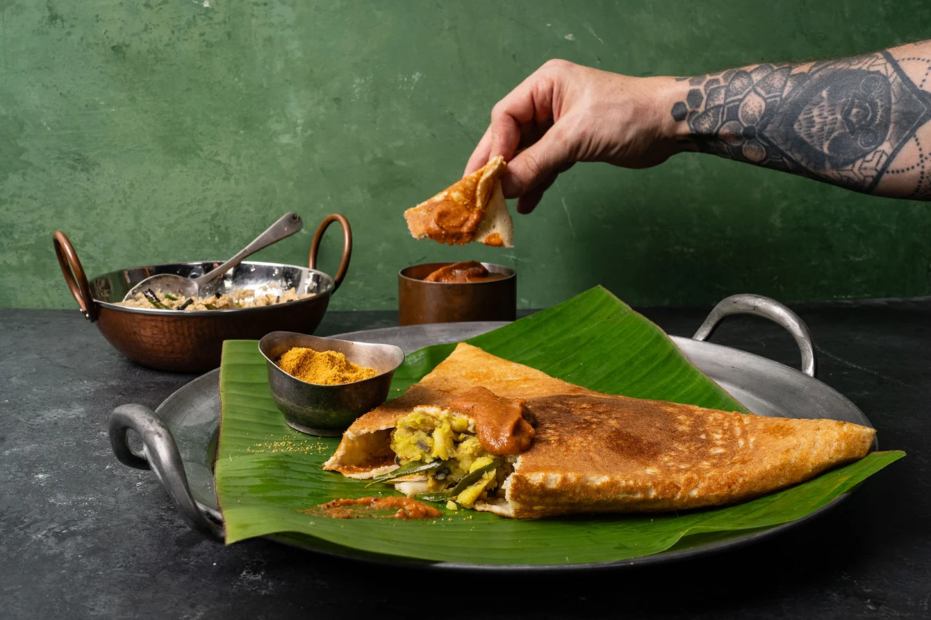 A plated masala dosa folded into a long triangle on a white plate with small bowls of coconut chutney and sambar<!-- inpage-recommendation:22359 --> on a wooden table, warm natural kitchen light