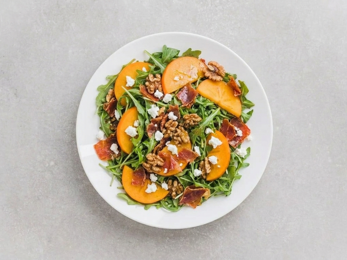 A plated persimmon (kaki) salad with arugula, crispy prosciutto shards, toasted walnuts, and crumbled goat cheese on a white plate