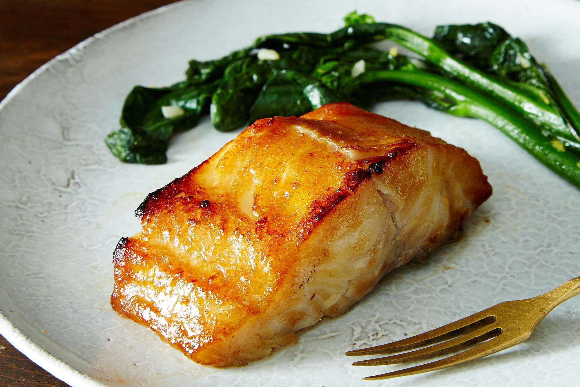 A plated piece of miso-glazed black cod with caramelized edges on a white plate, with steamed rice and sautéed bok choy on a warm kitchen table