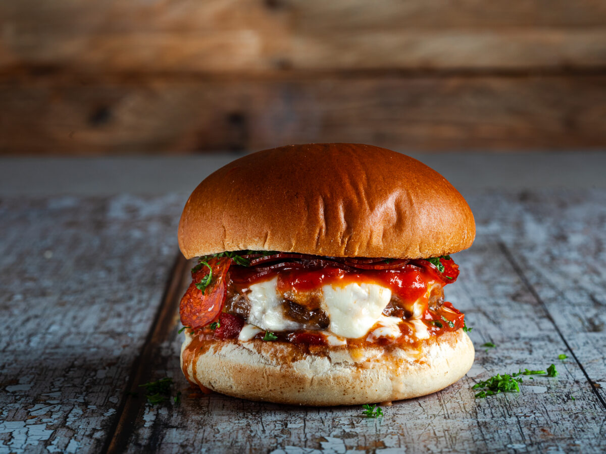 A plated pizza burger on a toasted bun with melted mozzarella, crisp pepperoni, and marinara sauce, set on a wooden table with a simple weeknight dinner setup
