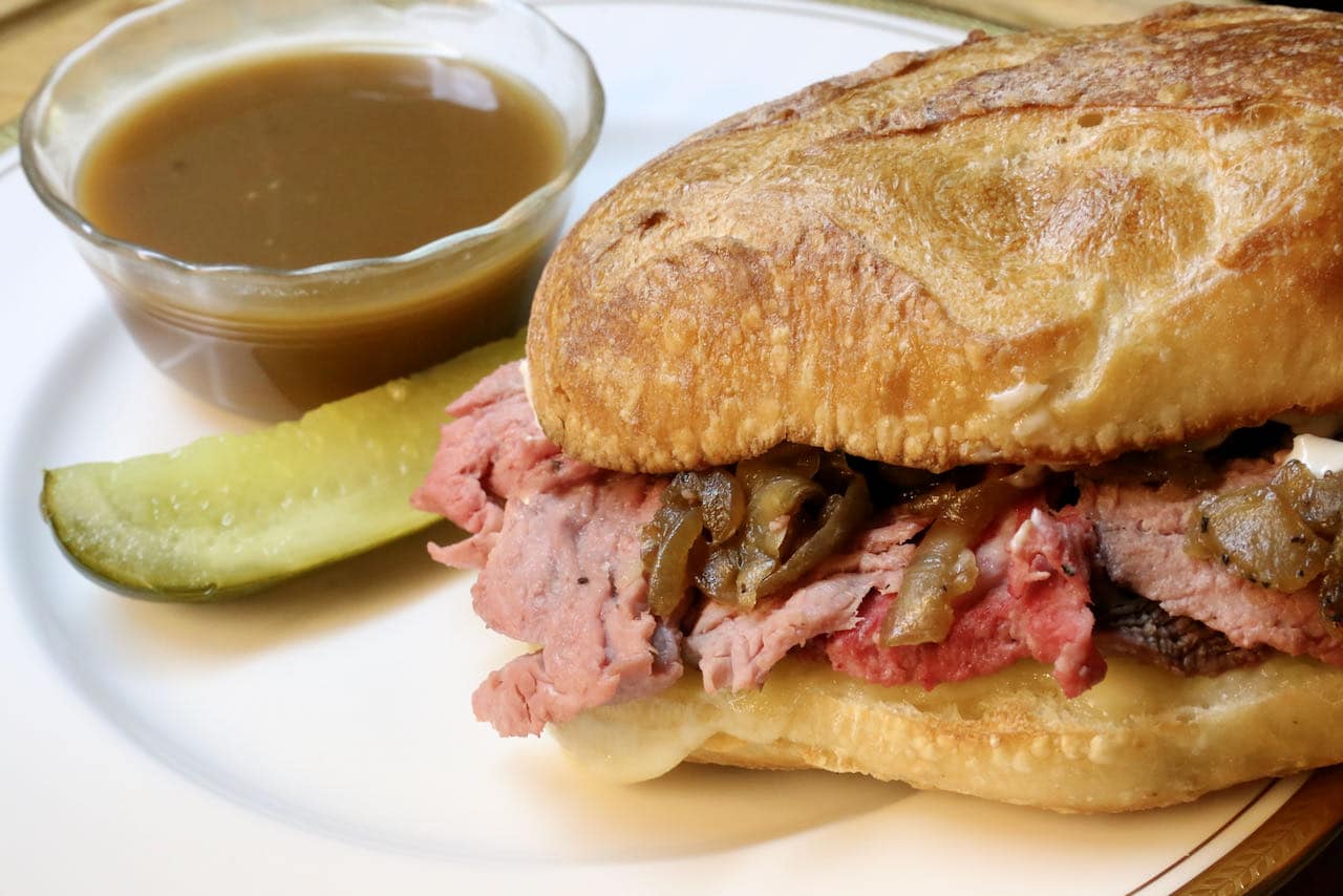 A plated prime rib French dip sandwich on a wooden cutting board with a small bowl of au jus for dipping, warm kitchen lighting