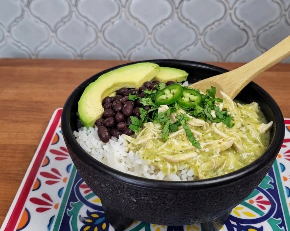 A plated rice bowl topped with creamy salsa verde shredded chicken, black beans, avocado slices, and cilantro on a dining table