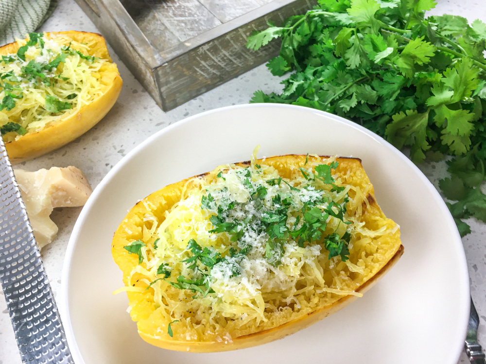 A plated roasted spaghetti squash half topped with garlic butter, parmesan, and chopped parsley on a simple white dinner plate on a warm kitchen table