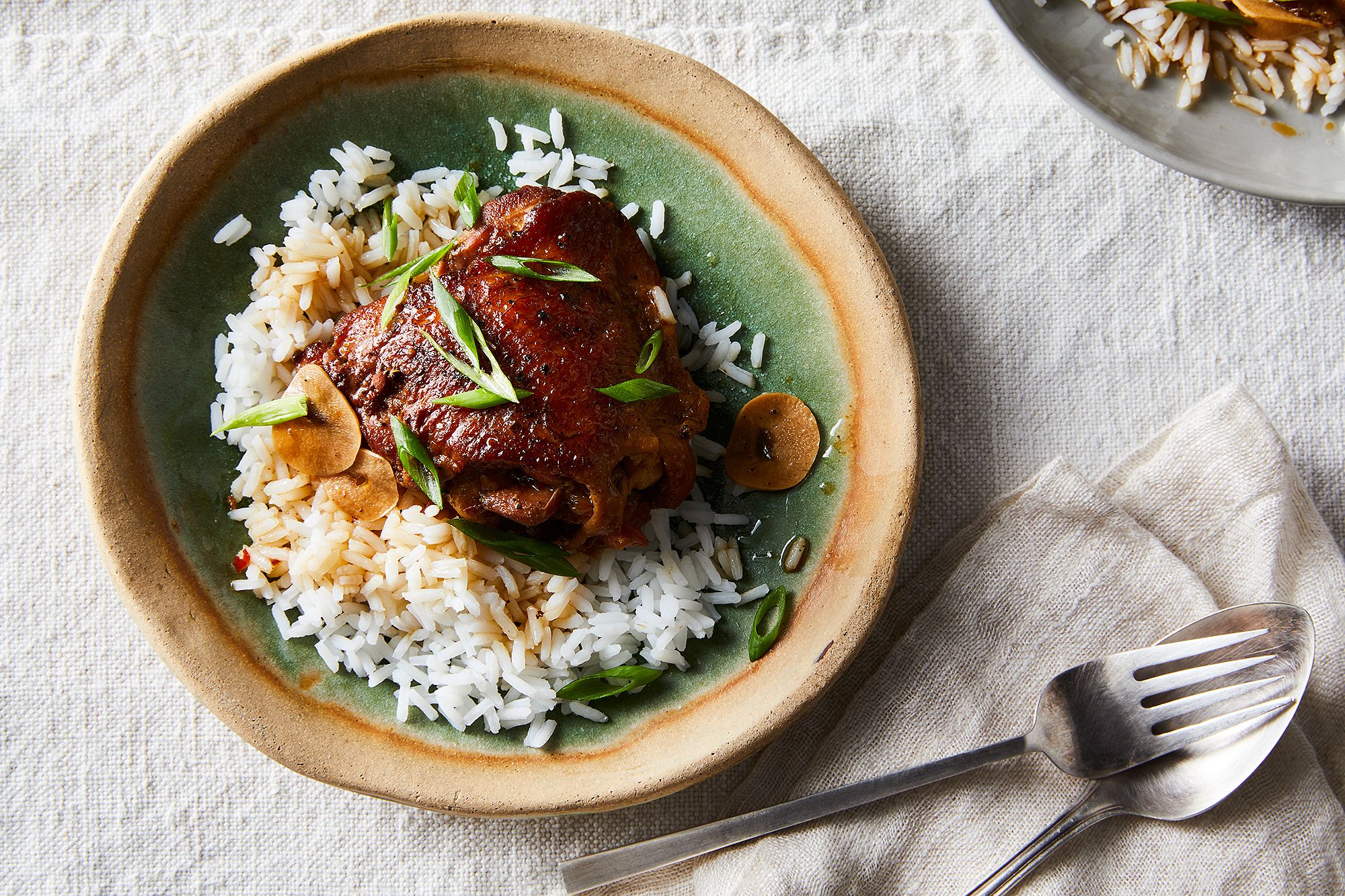 A plated serving of Filipino chicken adobo with glossy soy-vinegar sauce, steamed white rice, and a few sautéed greens on a simple ceramic plate on a warm kitchen table