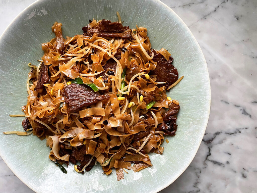 A plated serving of beef chow fun with glossy wide rice noodles, seared beef slices, bean sprouts, and scallions on a ceramic plate on a warm kitchen table