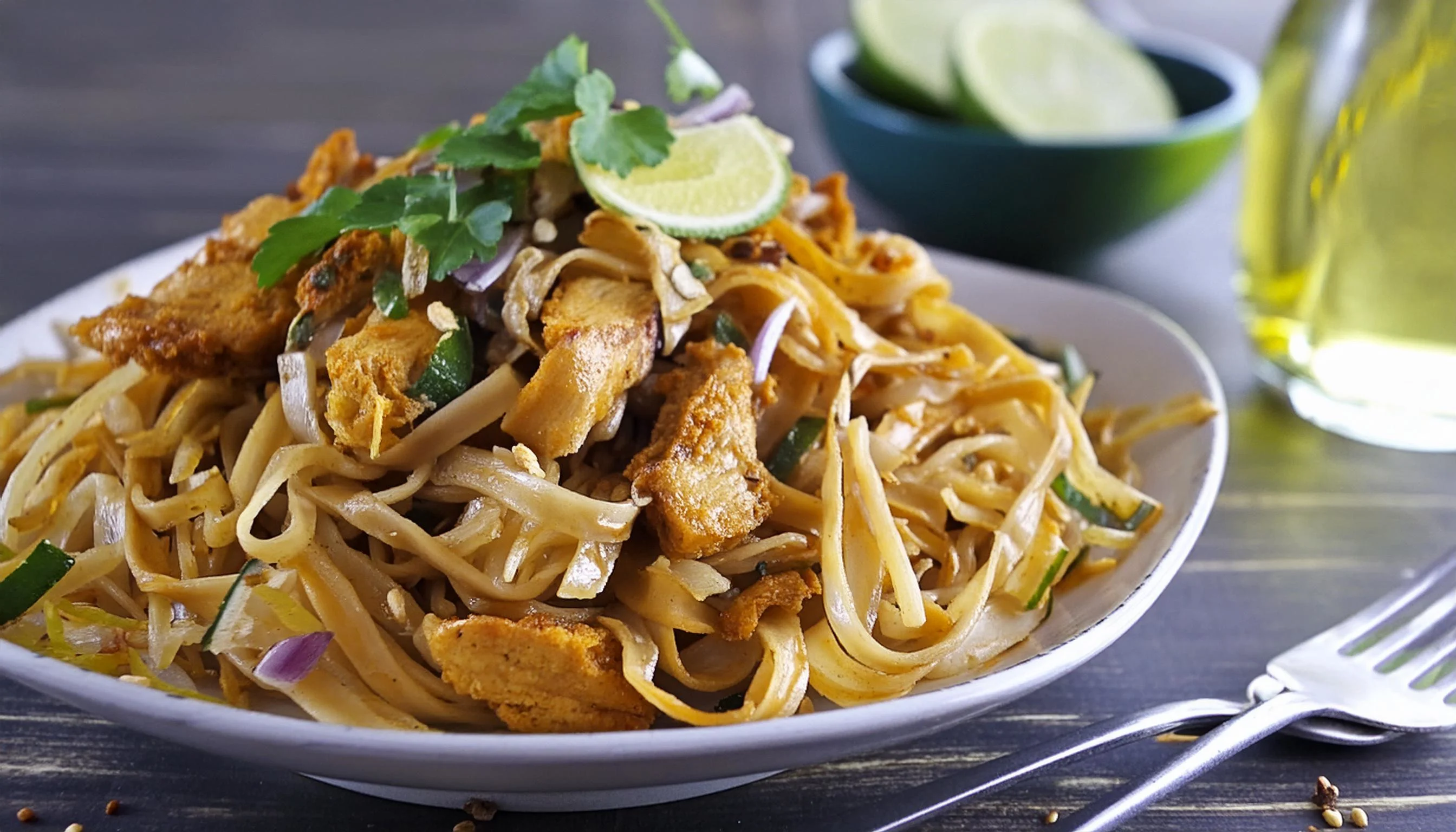 A plated serving of chicken pad thai with rice noodles, sliced chicken, bean sprouts, crushed peanuts, lime wedges, and green onions on a simple dinner table