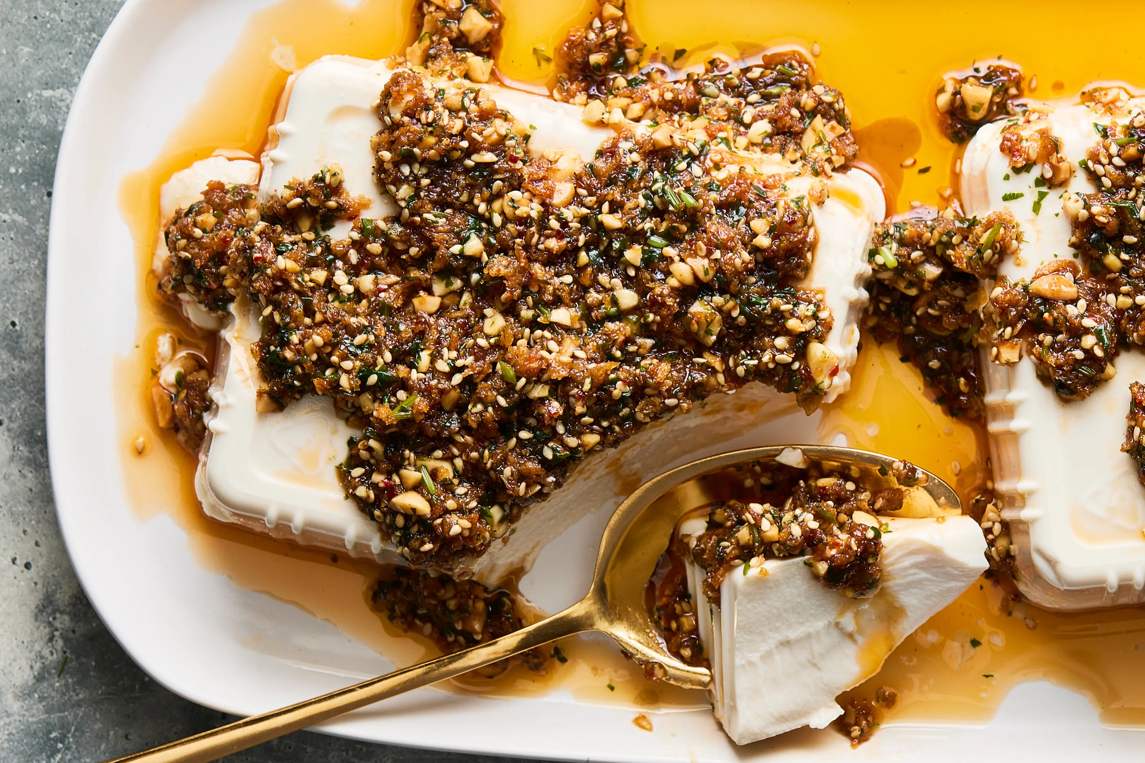 A plated serving of crispy-edged silken tofu coated in a glossy soy garlic glaze, topped with chili crisp, sliced scallions, and sesame seeds on a neutral ceramic plate on a warm-lit kitchen table