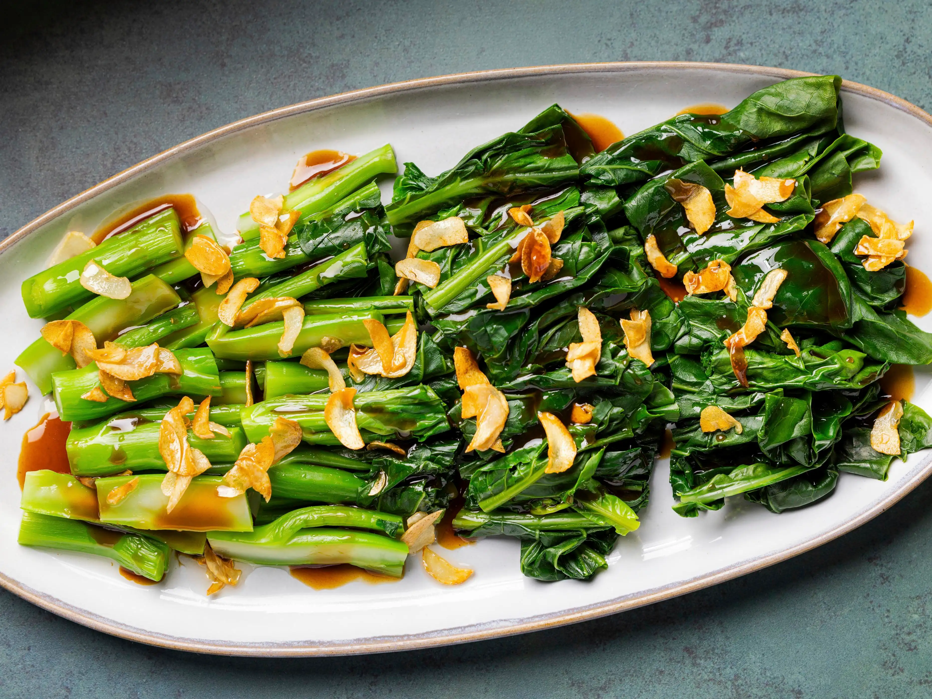 A plated serving of glossy gai lan (Chinese broccoli) with bright green leaves and tender stems coated in a garlic oyster sauce, set on a white plate on a warm kitchen table