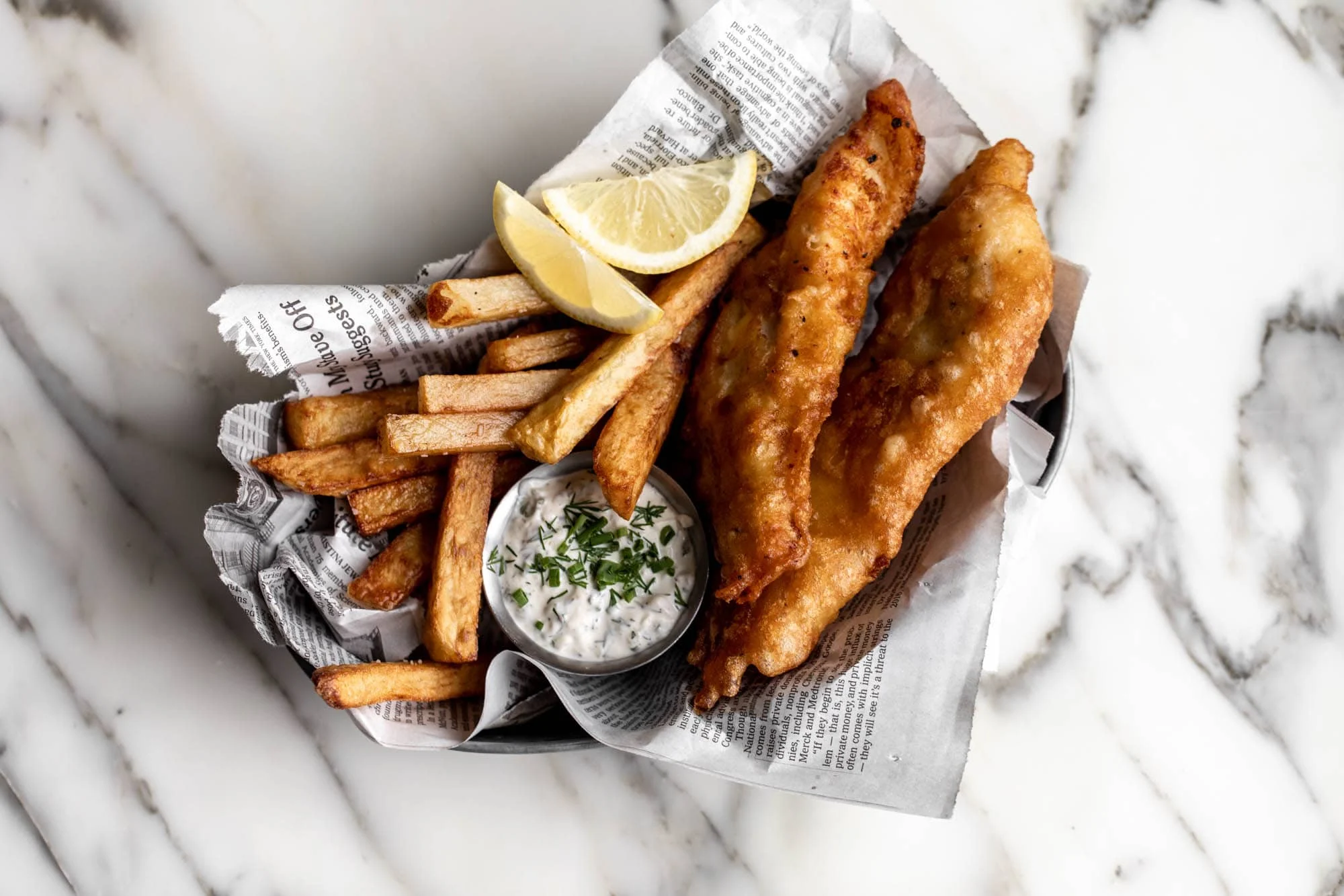 A plated serving of golden beer-battered fish with crispy fries, lemon wedges, and a small ramekin of tartar sauce on a wooden table in warm natural light