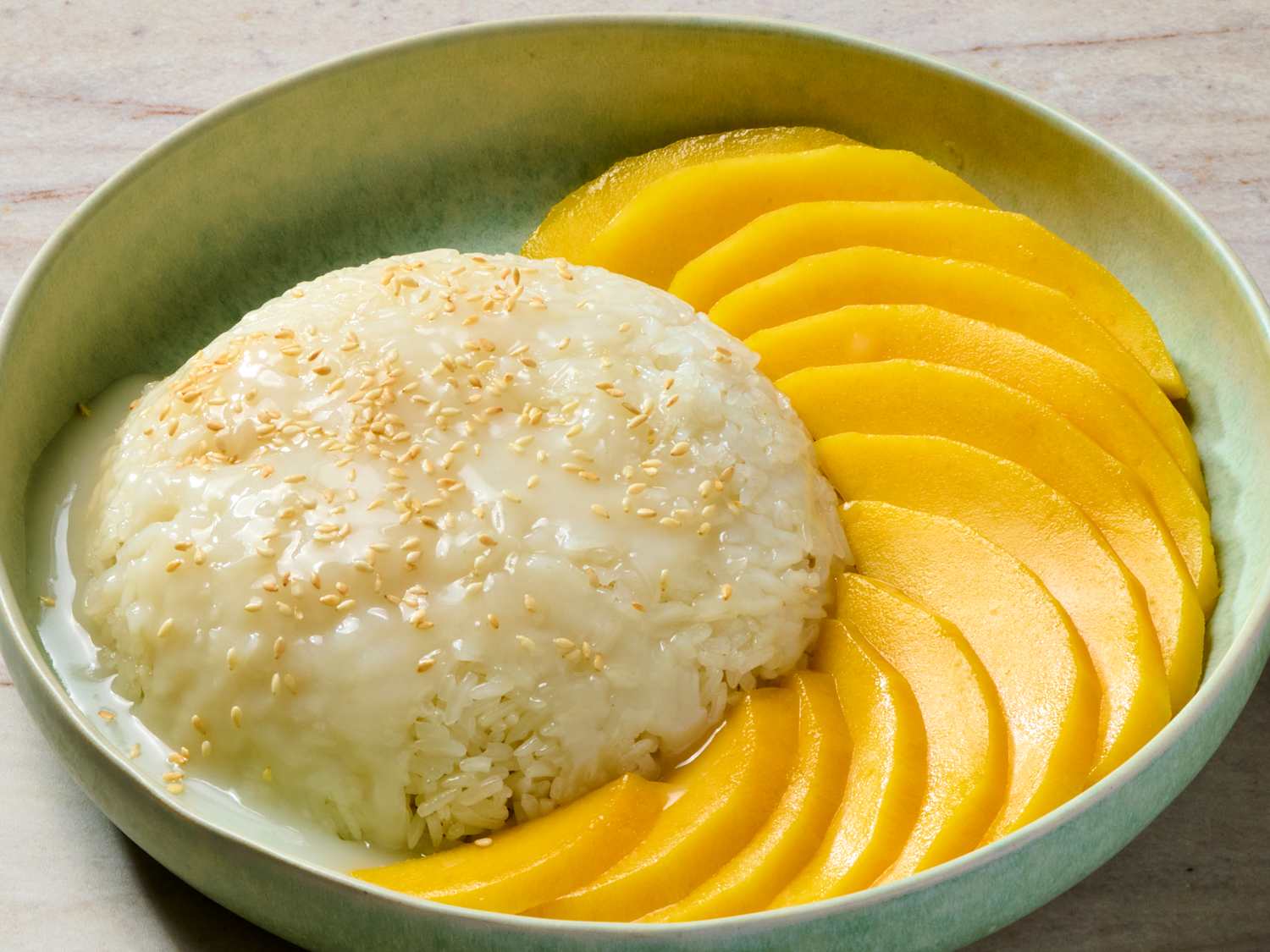 A plated serving of mango glutinous rice with glossy coconut sauce drizzled over sticky rice, fanned slices of ripe mango, and a small bowl of extra sauce on a warm-toned table setting