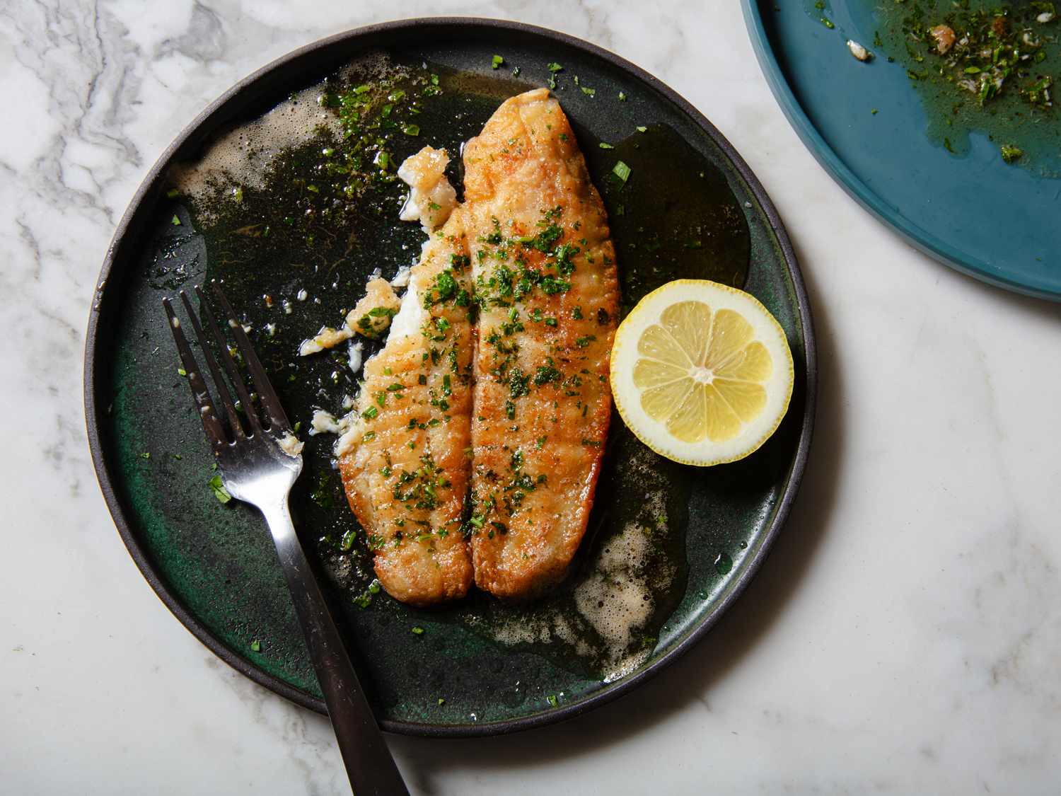 A plated sole meuniere fillet with browned butter sauce, lemon slices, and chopped parsley on a white plate on a softly lit kitchen table