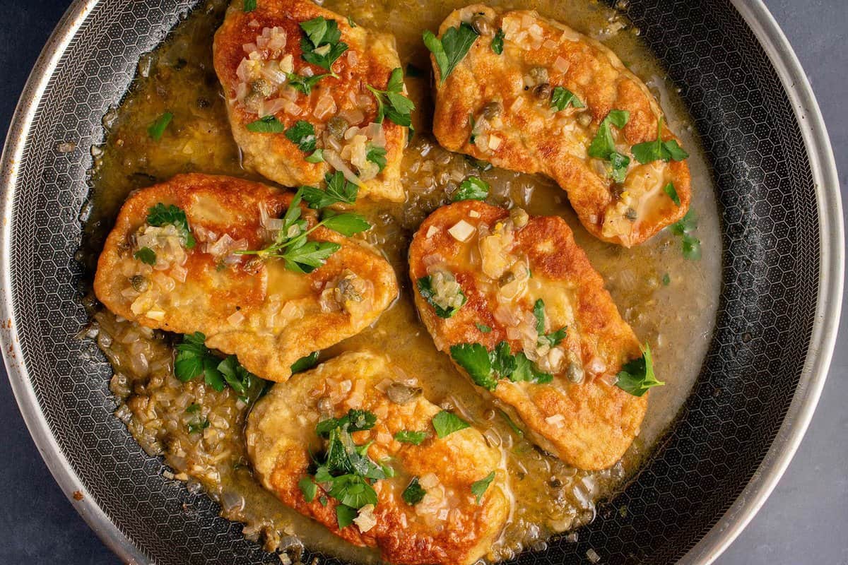 A plated stack of golden-brown crispy seitan cutlets drizzled with a glossy lemon-caper pan sauce on a white dinner plate, set on a warm kitchen table with soft natural light