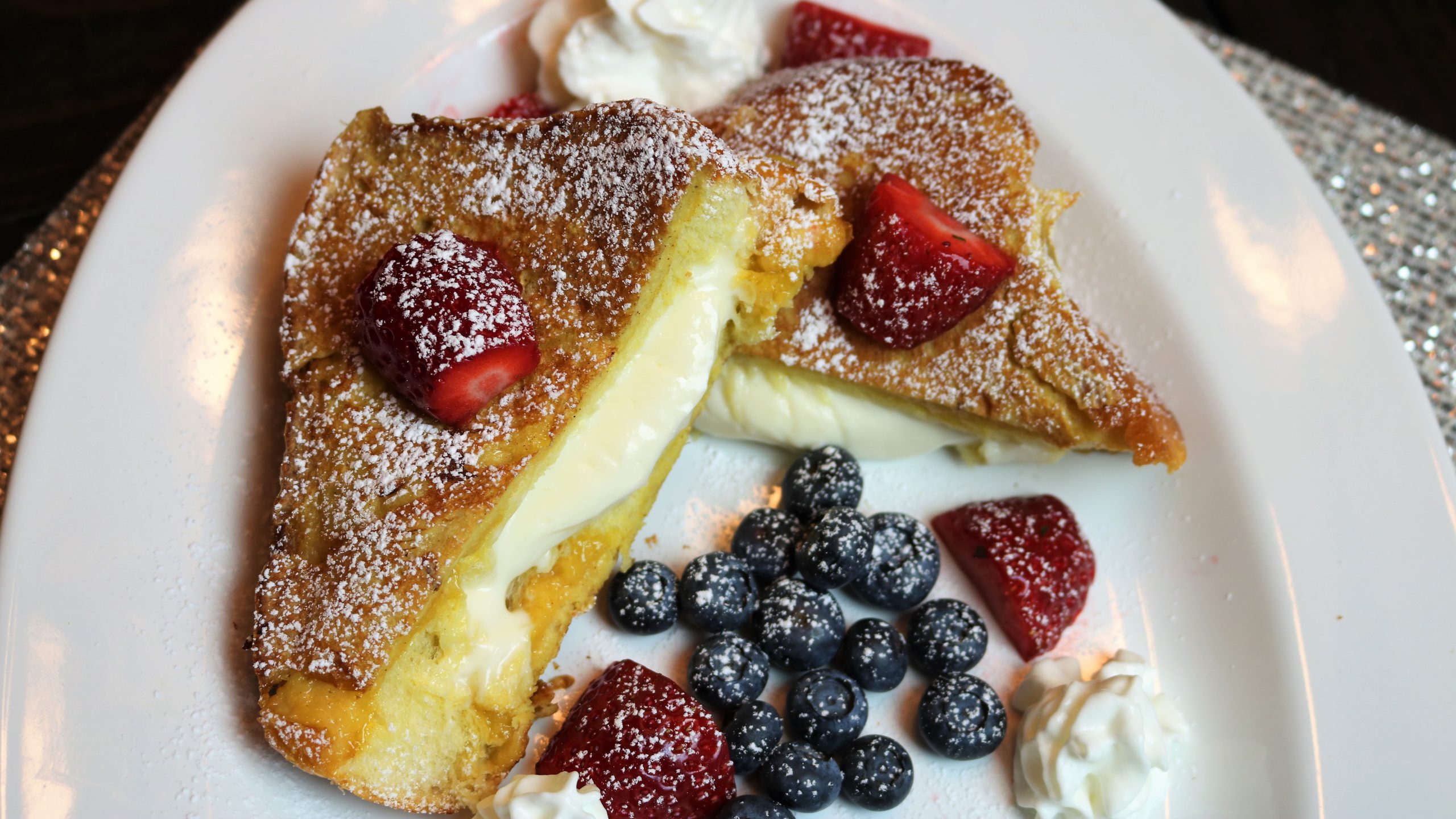 A plated stack of stuffed French toast cut open to show a creamy filling, topped with powdered sugar, fresh berries, and warm maple syrup on a sunlit kitchen table