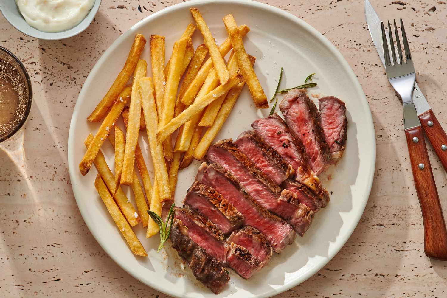 A plated steak frites dinner with sliced seared steak, a pile of crisp golden fries, and a small ramekin of Dijon shallot sauce on a simple stoneware plate