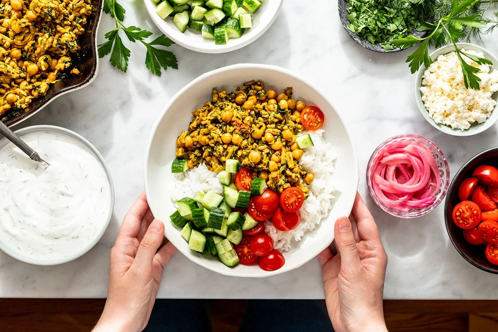 A plated vegetarian chickpea shawarma bowl with crispy spiced chickpeas, rice, sliced cucumbers, tomatoes, red onion, parsley, and a drizzle of lemon tahini sauce on a rustic dinner table