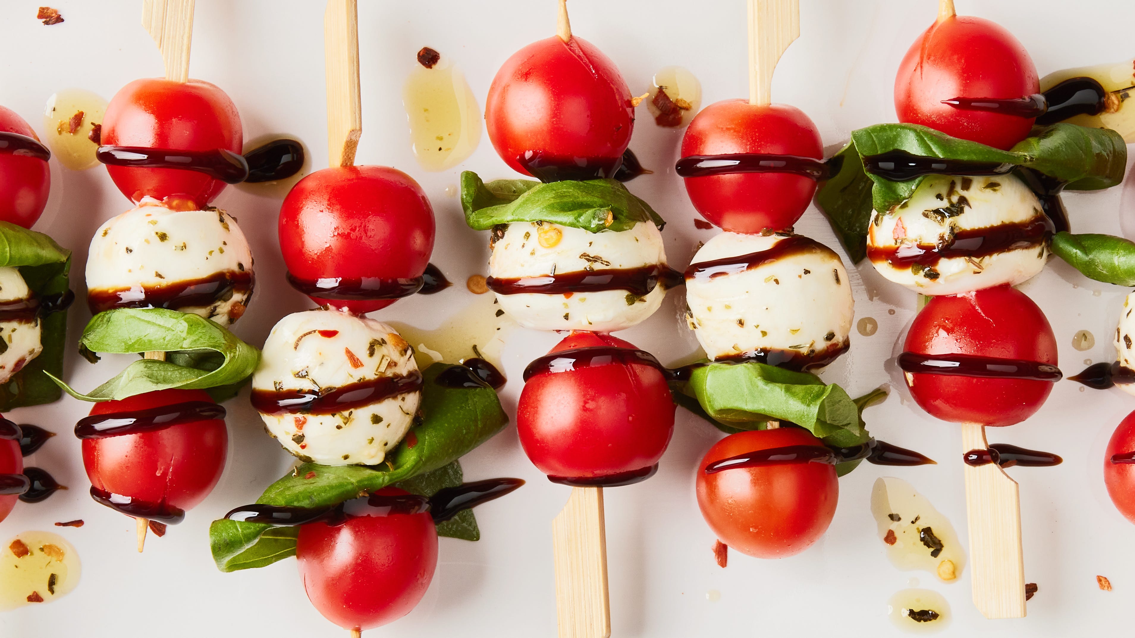 A platter of Caprese skewers with cherry tomatoes, fresh mozzarella balls, and basil leaves drizzled with balsamic glaze on a light kitchen counter
