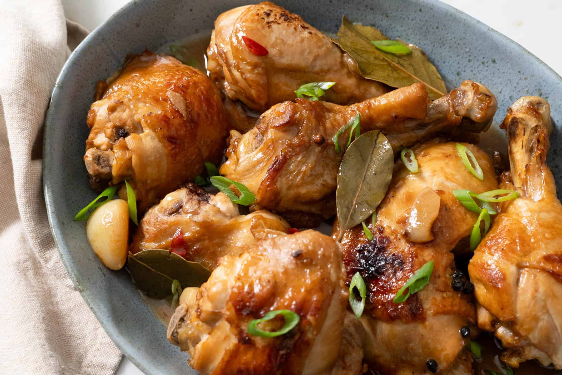 A platter of Estrada-style chicken adobo with browned chicken pieces in a glossy soy-vinegar sauce, scattered with fried garlic and sliced scallions, set on a wooden table with a bowl of steamed rice