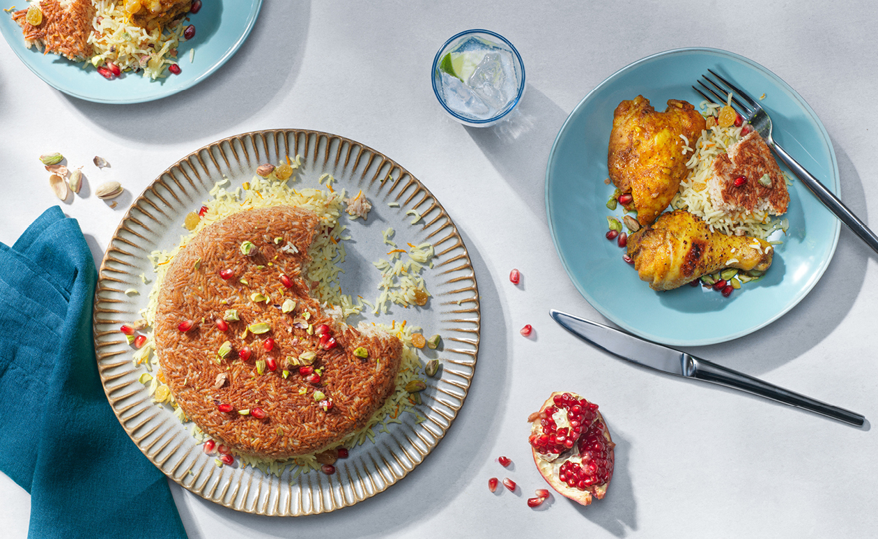 A platter of Persian-style saffron rice with a golden crispy tahdig crust, topped with tender chicken and scattered barberries, set on a simple dinner table with warm natural light