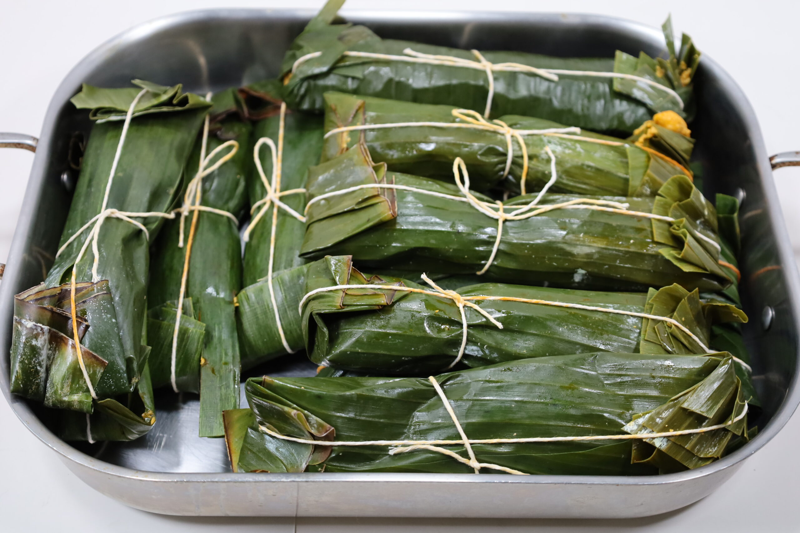 A platter of Puerto Rican pasteles wrapped in banana leaves with kitchen twine, one pastel opened to show the savory pork filling, on a warm-toned kitchen table
