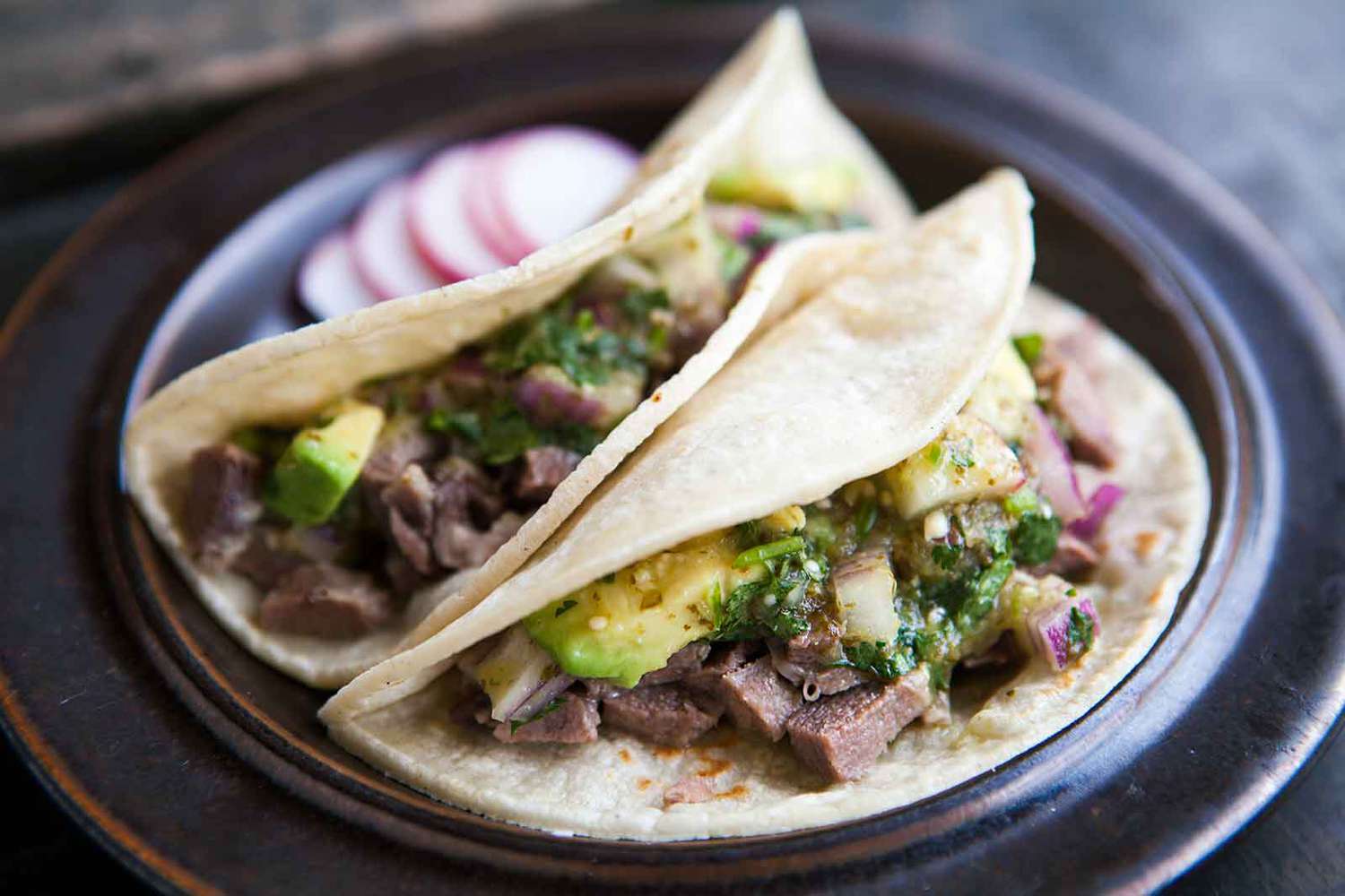 A platter of beef tongue tacos with crisped slices of lengua, salsa verde, diced onion, cilantro, and lime wedges on a wooden table