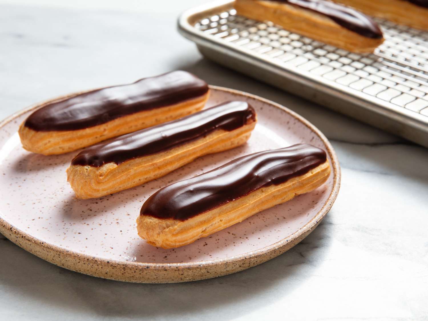 A platter of classic chocolate eclairs with glossy chocolate glaze, split open to show vanilla pastry cream, on a bright kitchen table