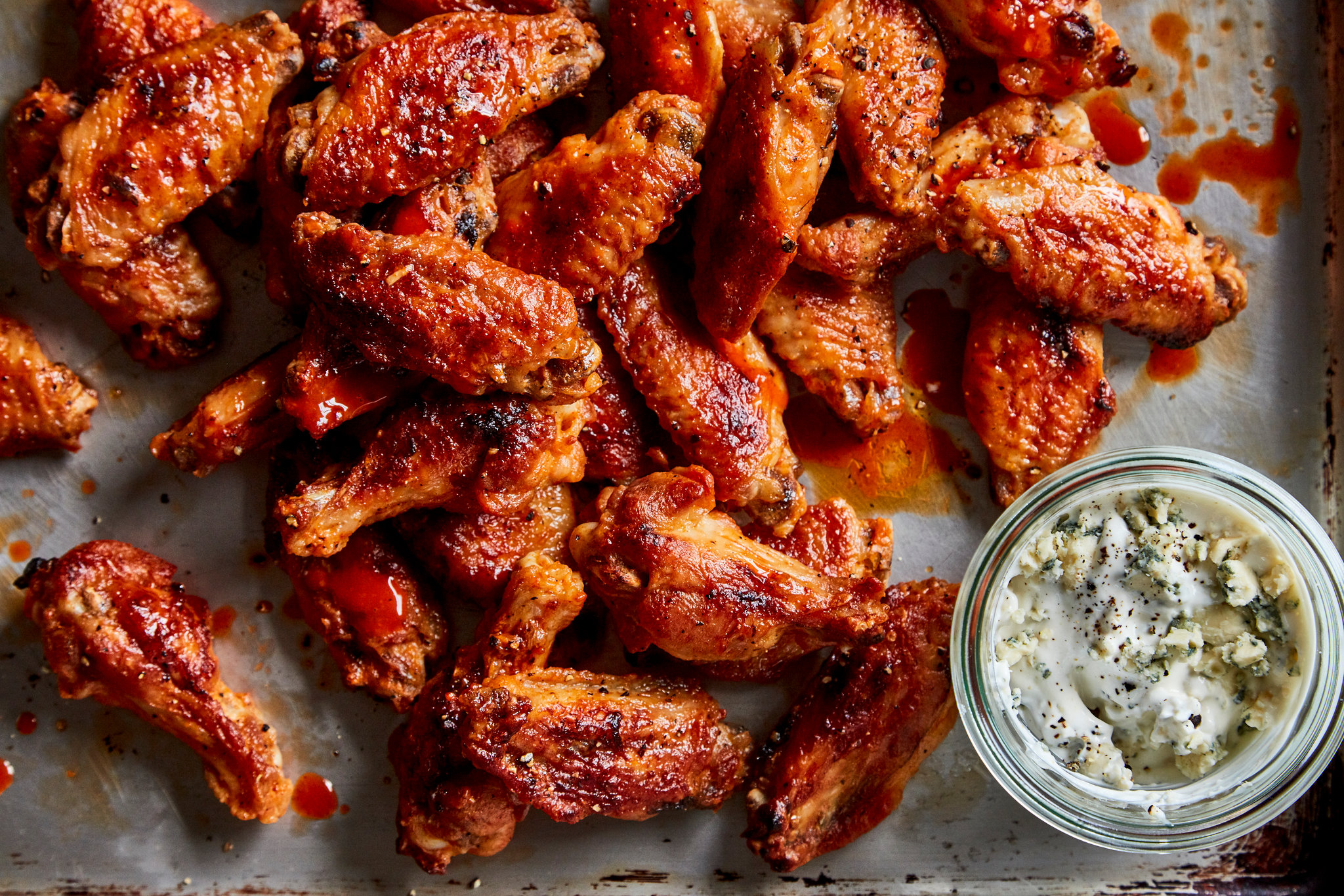 A platter of crispy Buffalo hot wings glazed in a glossy orange-red sauce with celery sticks and a small bowl of ranch on a wooden table
