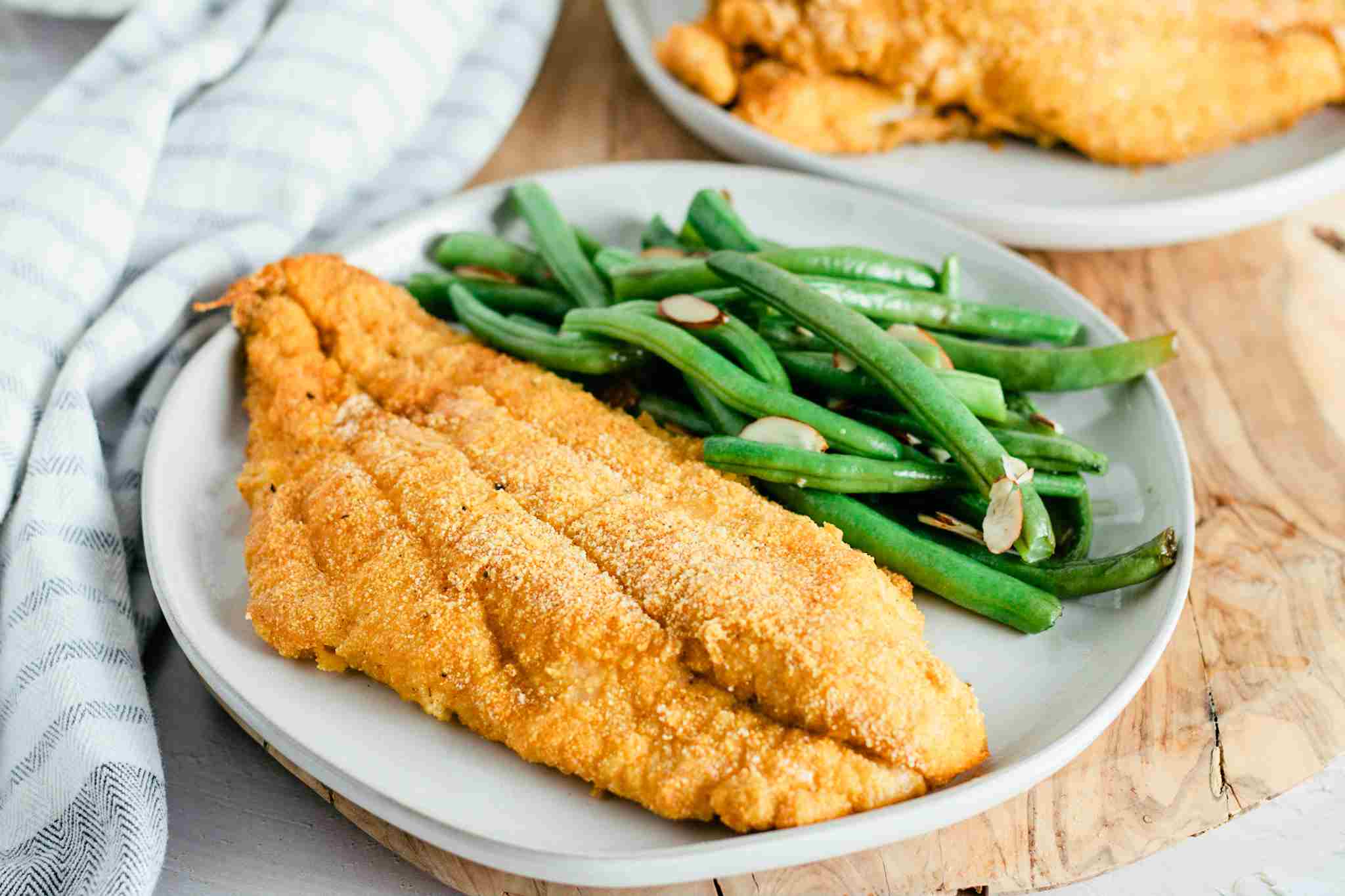 A platter of crispy baked catfish fillets with a golden cornmeal crust, lemon wedges, and chopped parsley on a warm-toned wooden table