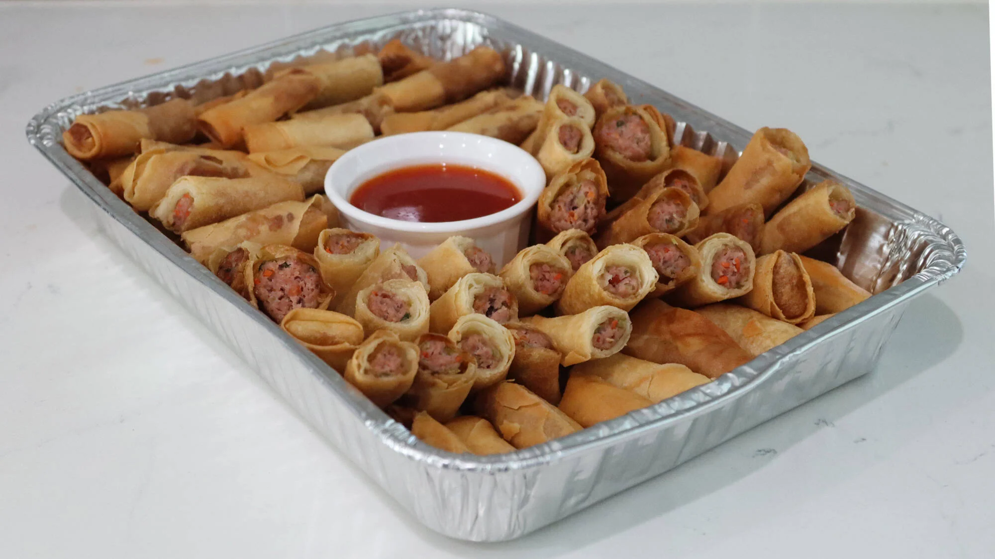 A platter of freshly fried Filipino pork lumpia cut on a diagonal, stacked with a small bowl of sweet chili dipping sauce on a wooden table with warm kitchen lighting