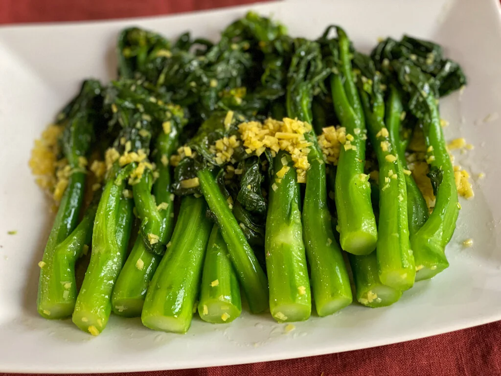 A platter of glossy garlic-ginger Chinese broccoli (gai lan) with bright green leaves and crisp stems, sprinkled with toasted sesame seeds on a simple dinner table