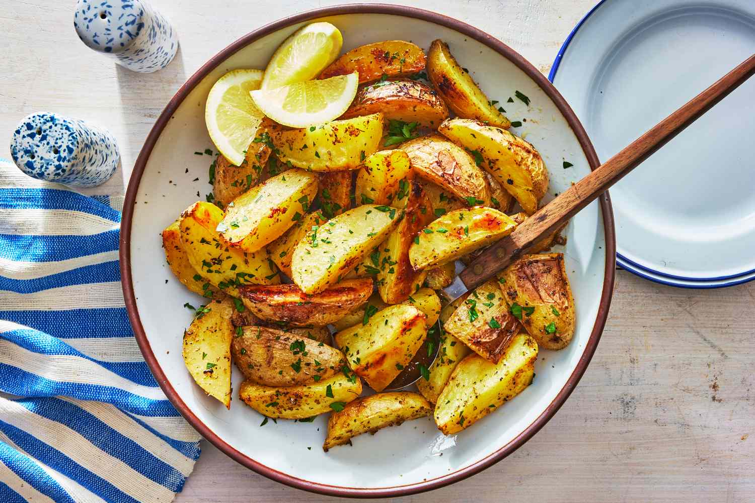 A platter of golden Greek lemon potatoes with crisp edges, scattered oregano and parsley, with lemon wedges on a rustic wooden table