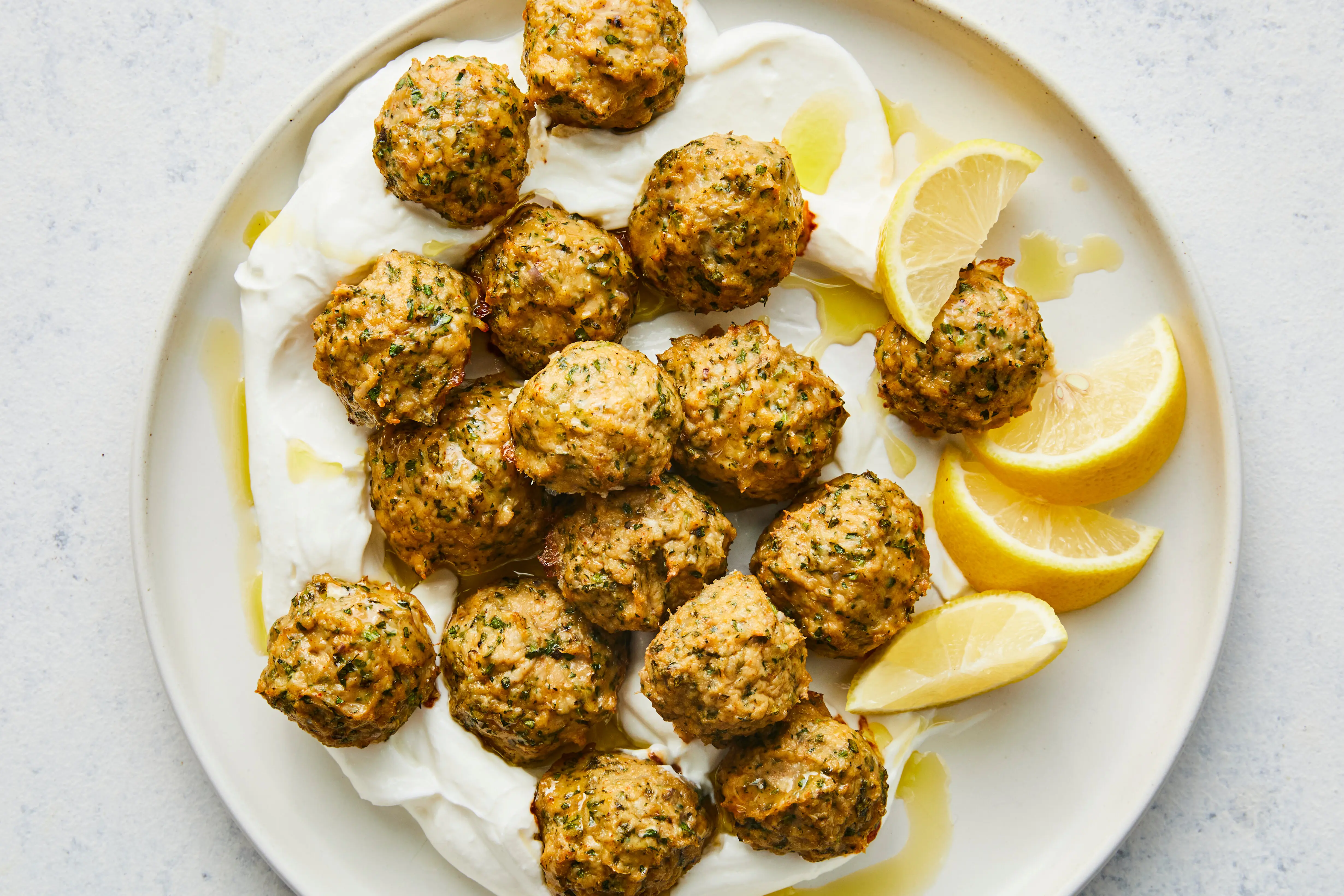 A platter of golden-brown Greek meatballs garnished with parsley and lemon wedges, with a small bowl of creamy lemon garlic yogurt sauce on a rustic wooden table