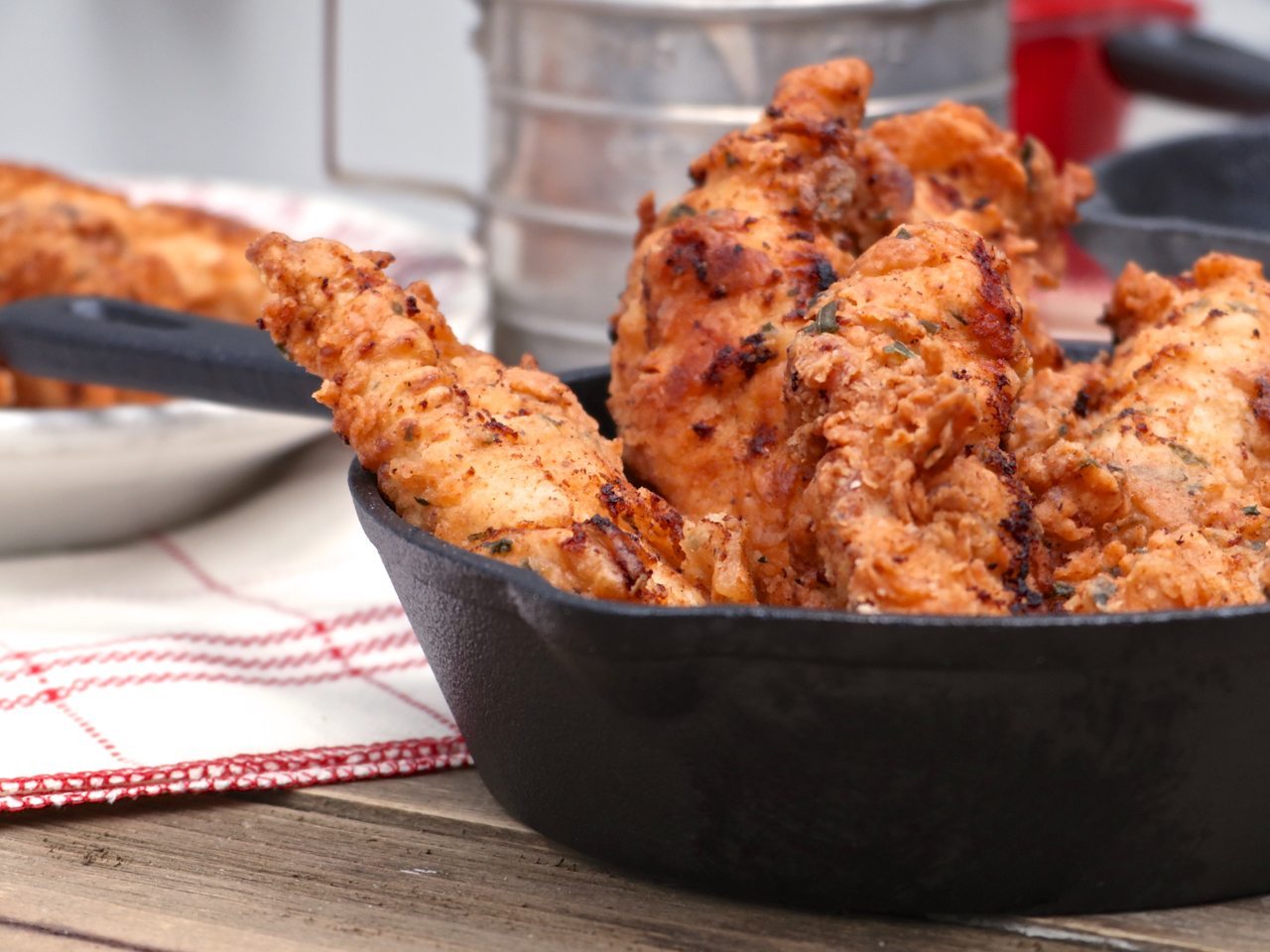 A platter of golden brown buttermilk fried chicken tenders with a craggy crisp coating, served with a small bowl of dipping sauce on a rustic kitchen table