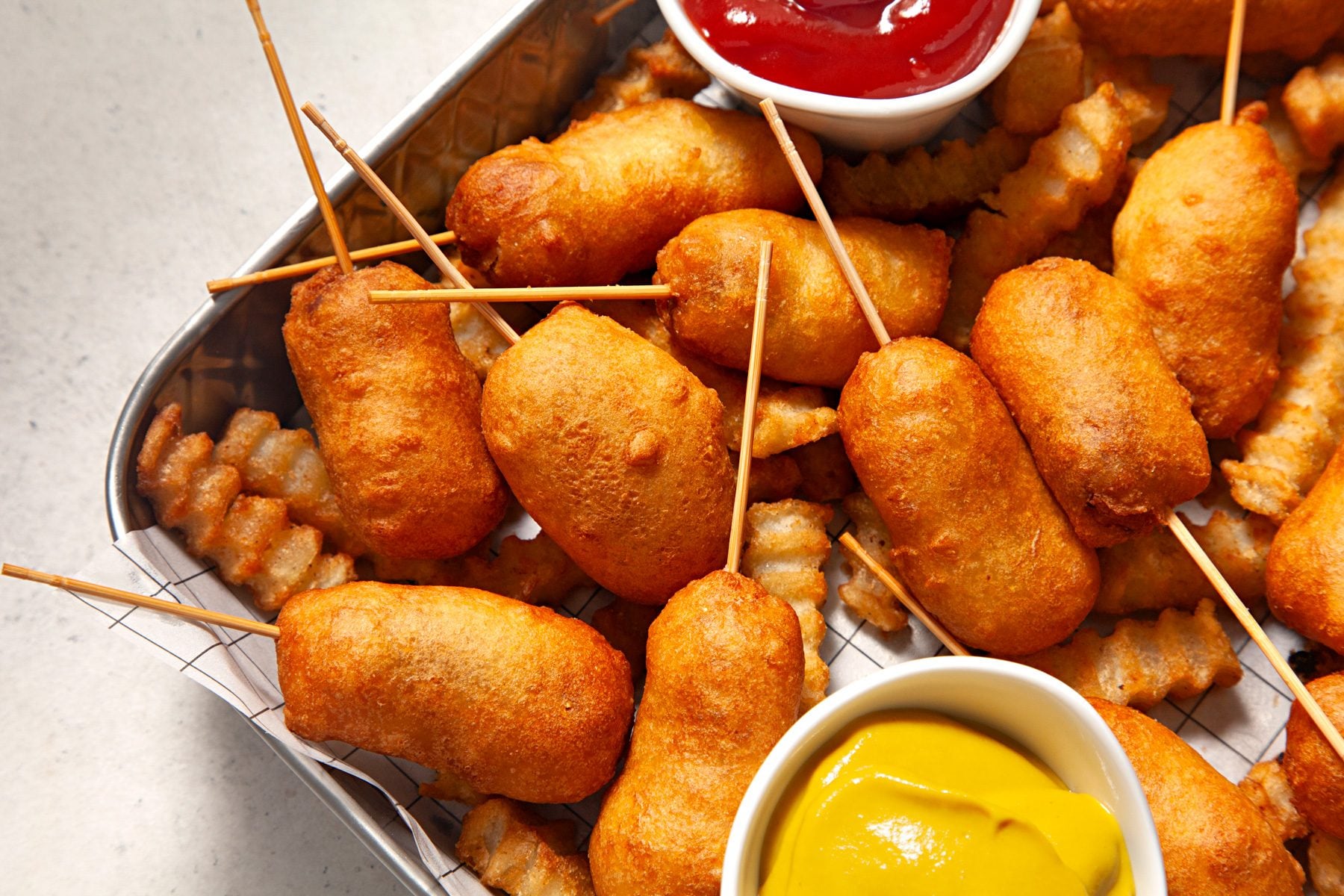 A platter of golden brown homemade pogos with crisp cornmeal coating, wooden sticks, and small bowls of ketchup and mustard on a warm kitchen table