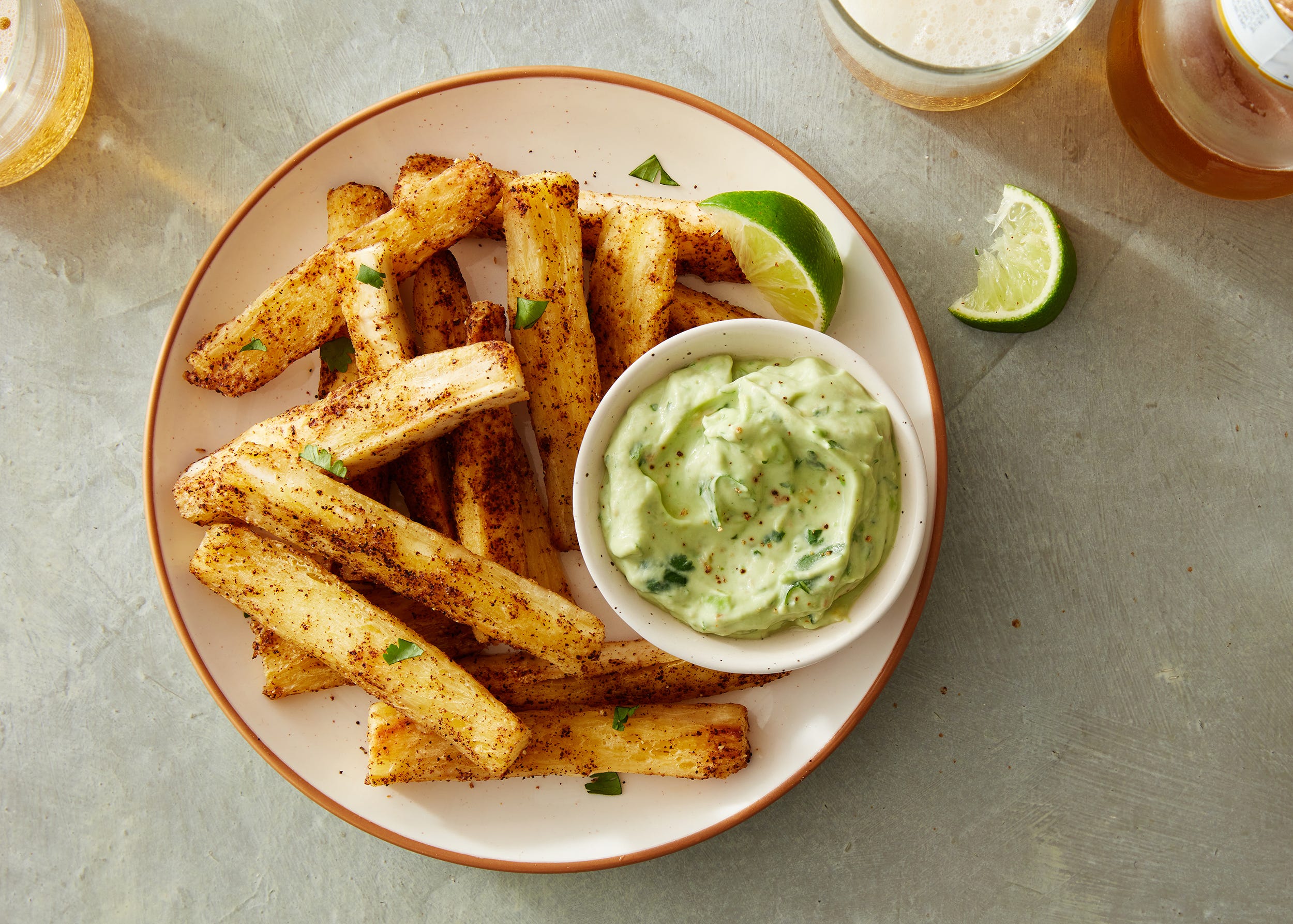 A platter of golden cassava (yuca) fries with a small bowl of creamy garlic lime aioli on a rustic kitchen table, warm natural light