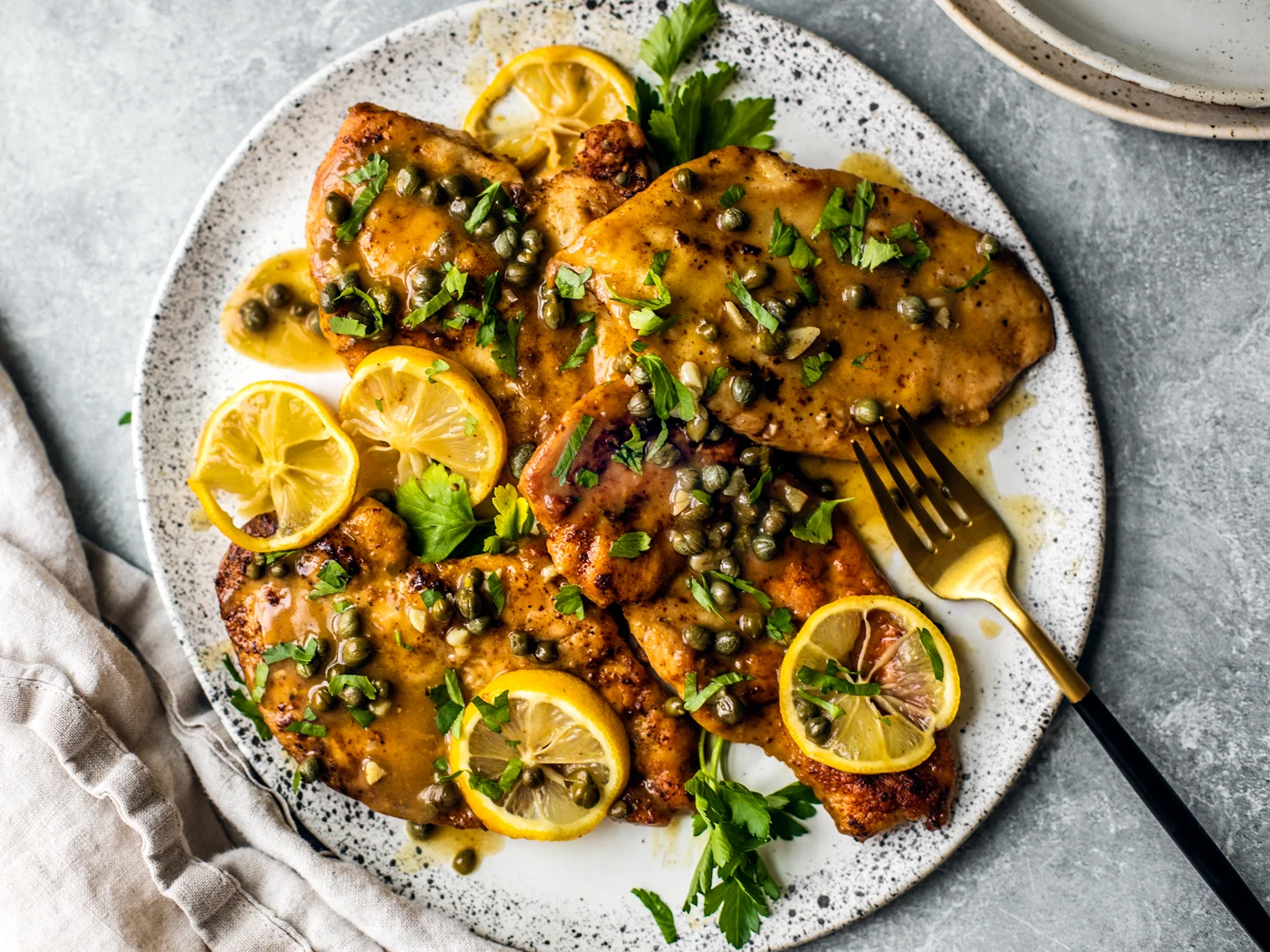 A platter of golden chicken piccata topped with capers and lemon slices, with a glossy butter sauce pooling around the chicken on a white serving dish on a warm kitchen table