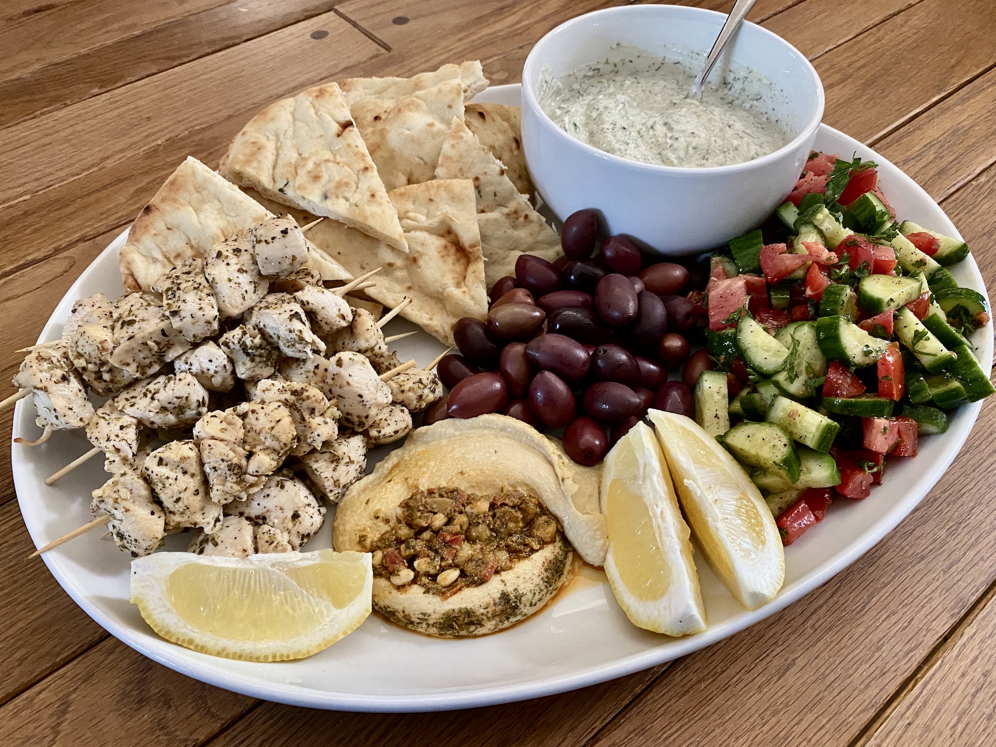 A platter of grilled Greek chicken souvlaki skewers with charred edges, warm pita, lemon wedges, sliced cucumber, and a bowl of creamy yogurt sauce on a bright kitchen table