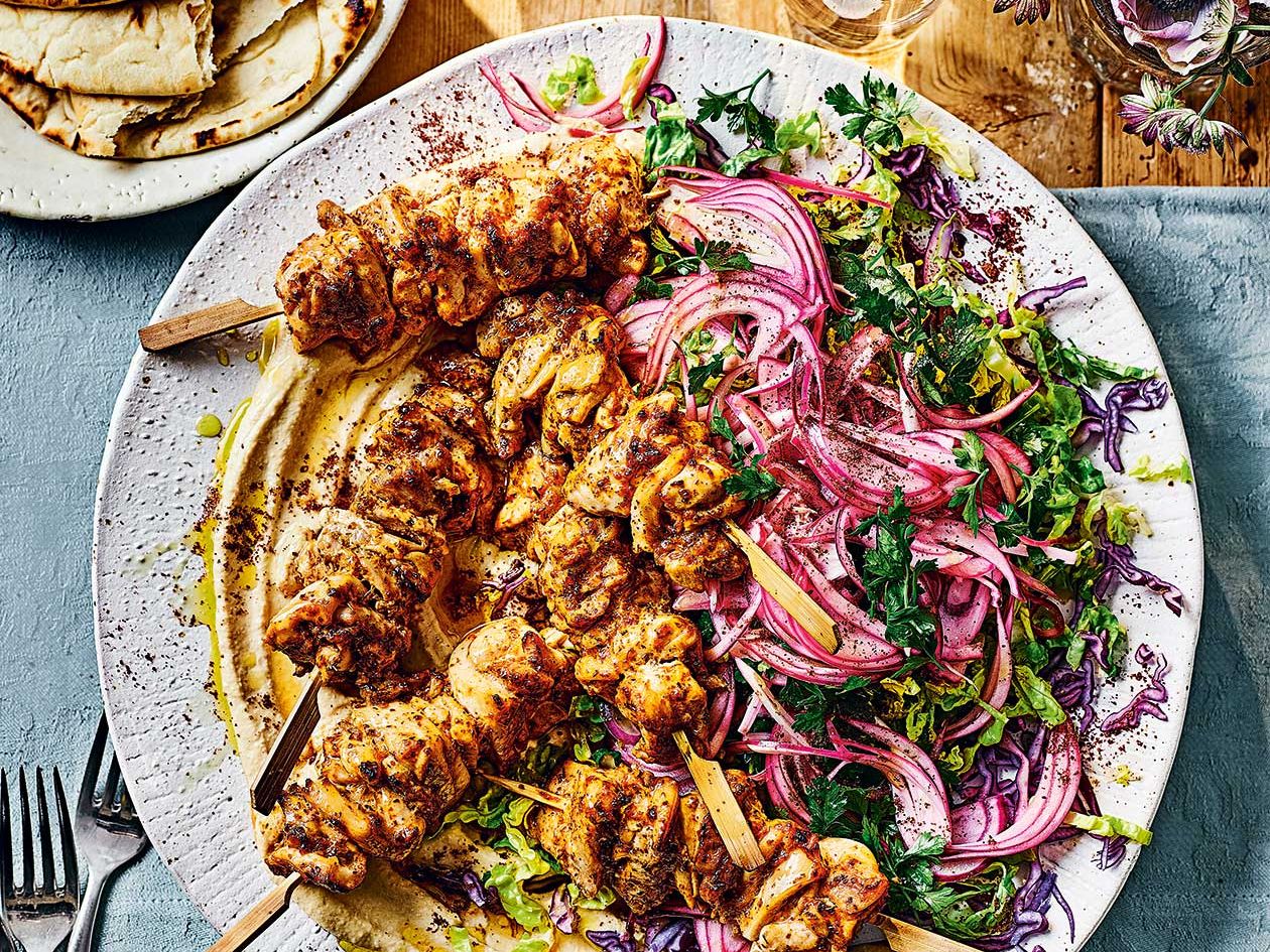 A platter of grilled chicken kabab skewers with charred edges, lemon wedges, and a small bowl of yogurt sauce on a wooden table