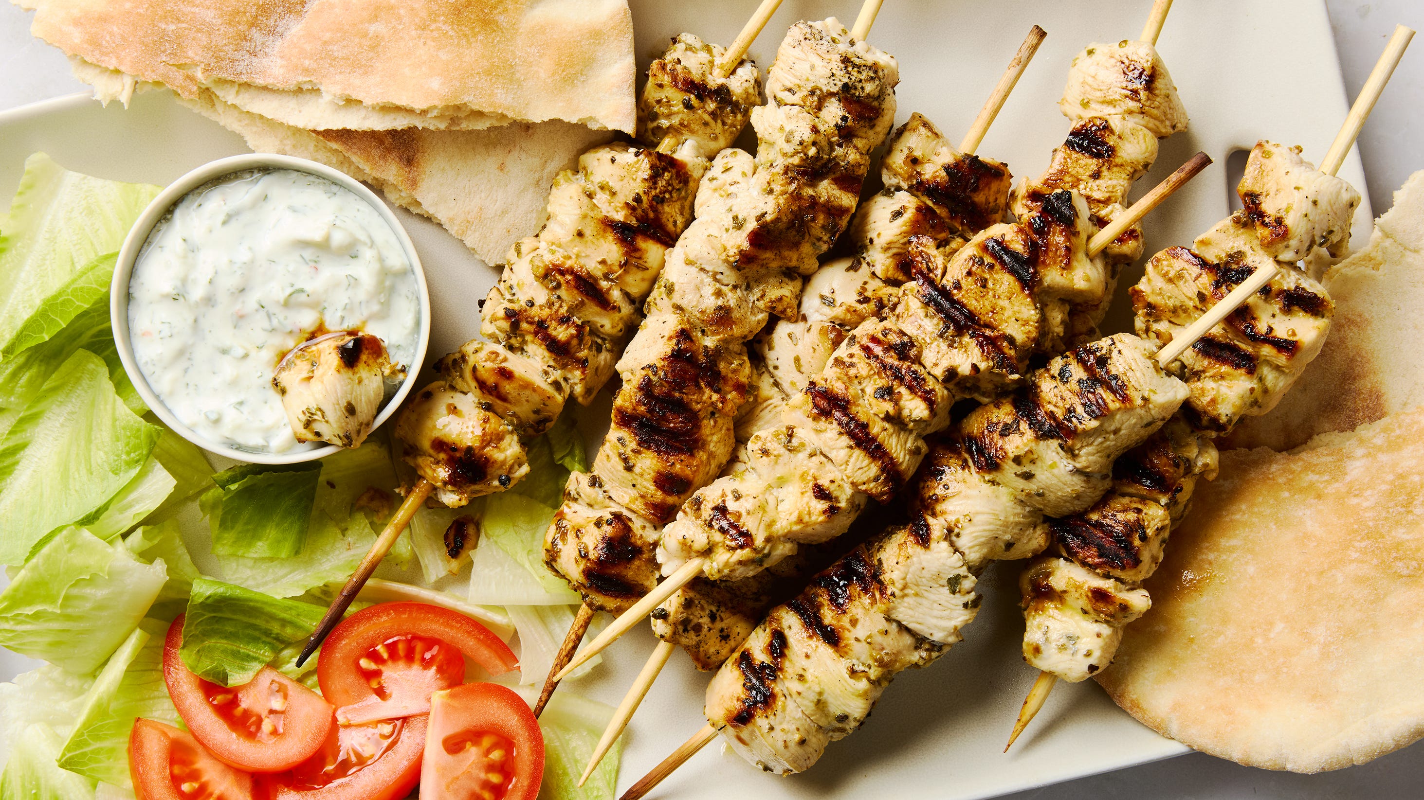 A platter of grilled chicken souvlaki skewers with charred edges, lemon wedges, sliced cucumbers, and warm pita on a wooden table in soft natural light