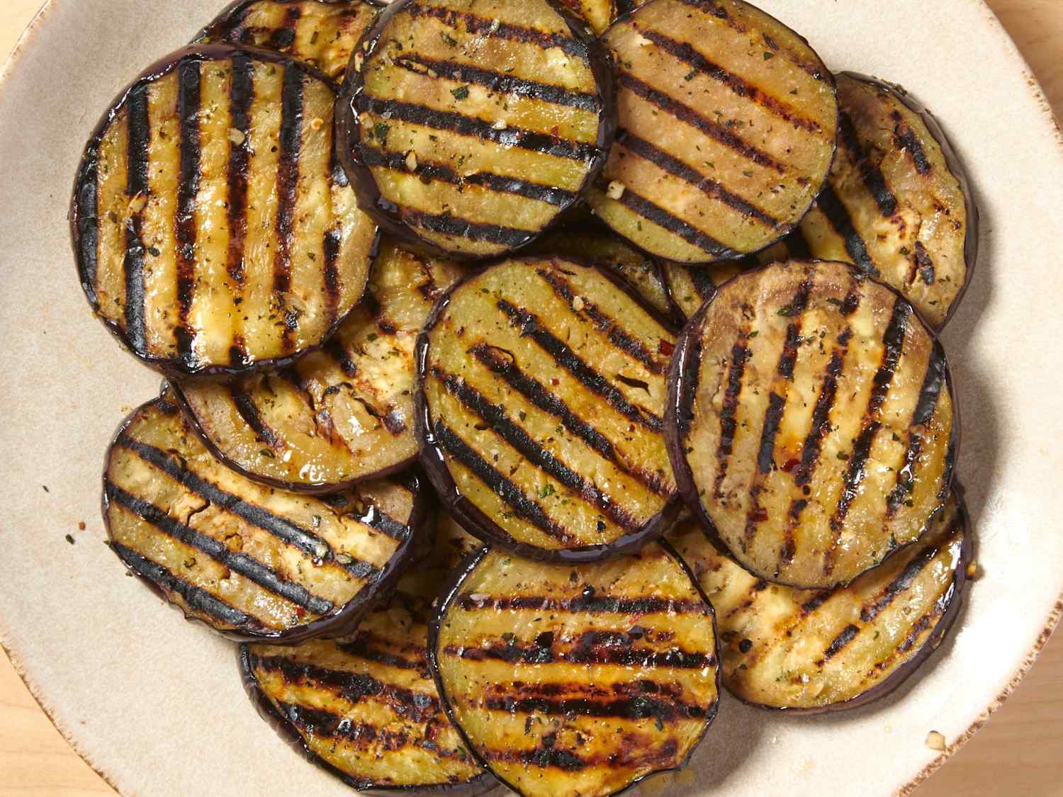 A platter of grilled eggplant slices with deep grill marks, glistening with olive oil and dotted with chopped herbs on a wooden table