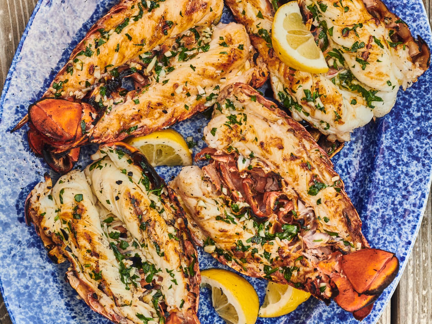 A platter of grilled lobster tails with charred edges, brushed with garlic herb butter and served with lemon wedges on a wooden table