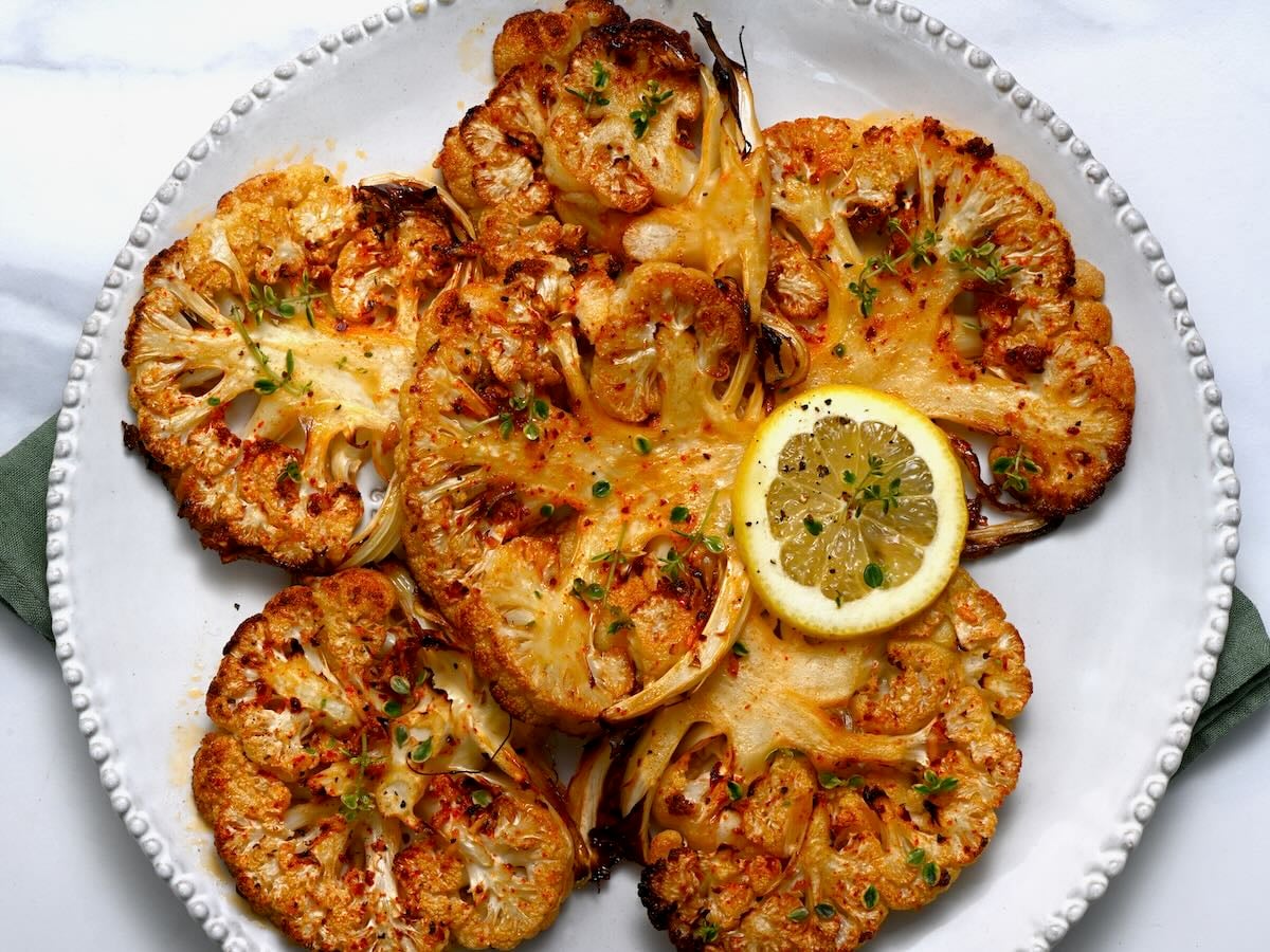 A platter of roasted cauliflower steaks with caramelized edges, spooned with lemon-garlic herb sauce, set on a simple wooden table with soft natural light
