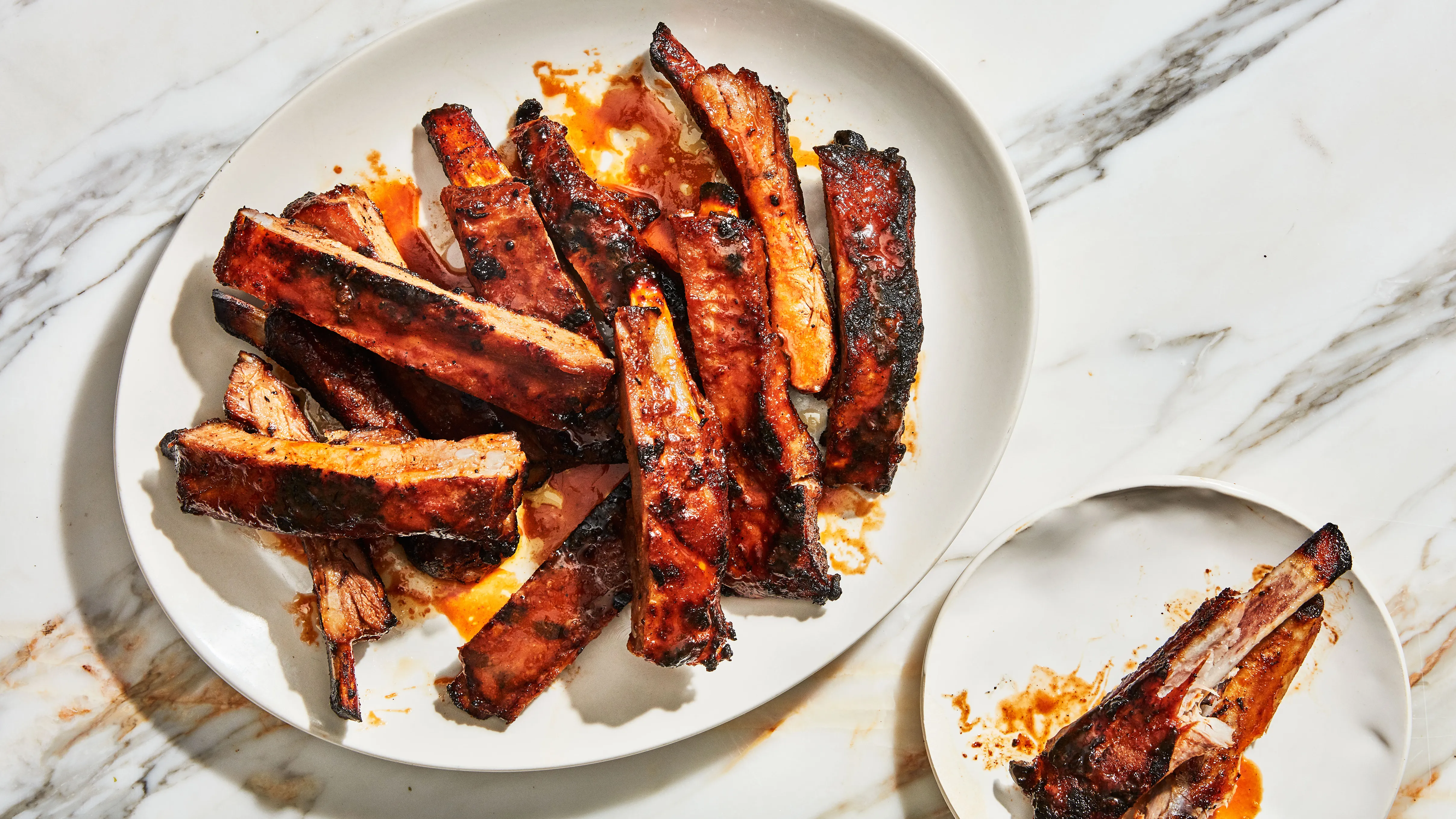 A platter of saucy pork ribs with caramelized edges, sliced into individual bones, on a wooden table with a small bowl of extra barbecue sauce and a simple dinner setting