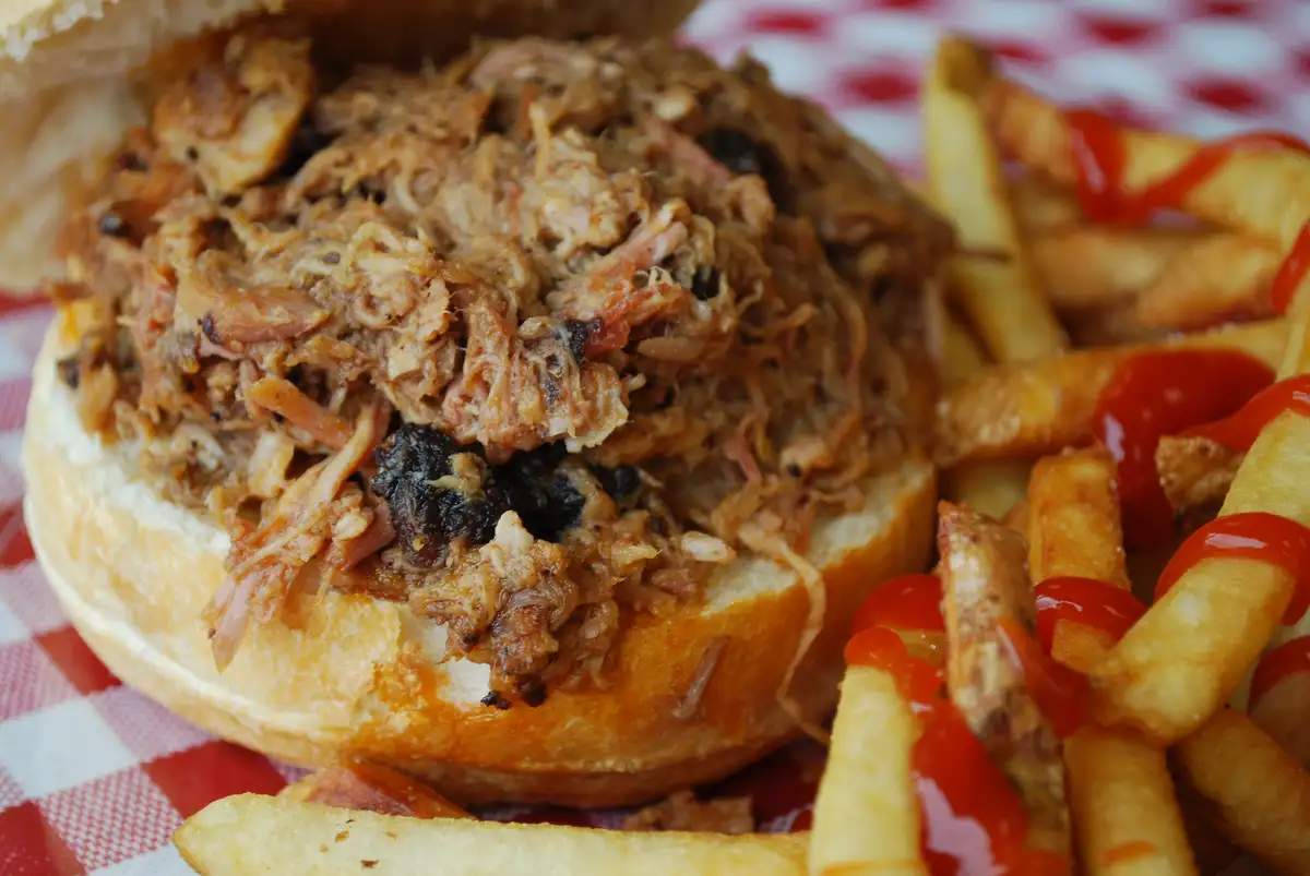 A platter of saucy pulled pork piled high with soft buns, pickles, and coleslaw on a warm kitchen table with natural window light