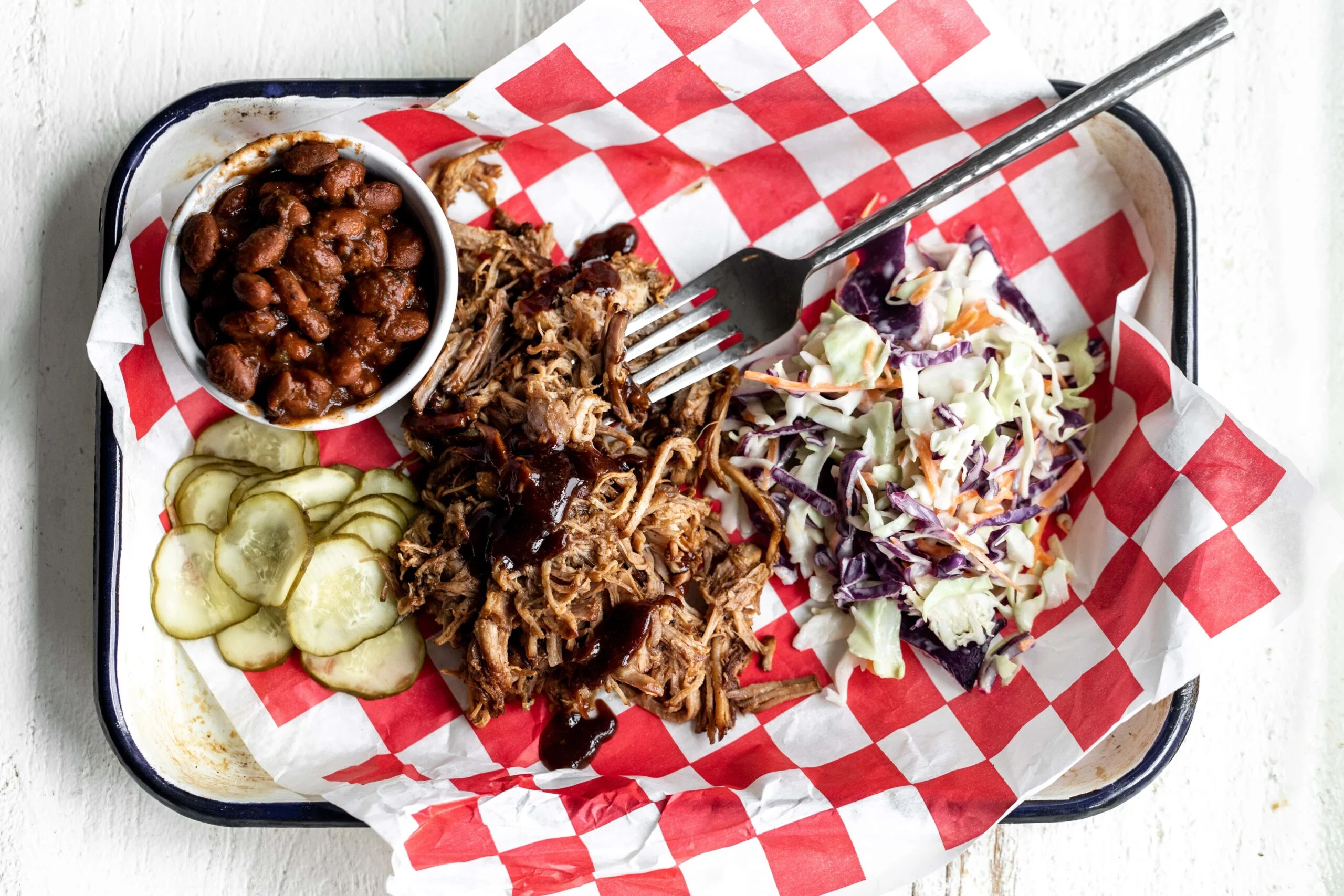 A platter of saucy pulled pork with a pile of shredded meat, pickles, and soft buns on a wooden table under warm kitchen lighting