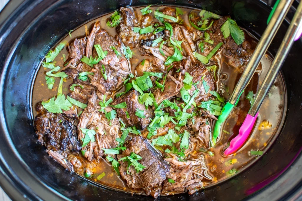 A platter of shredded beef barbacoa with crispy edges, served with warm corn tortillas, diced onion, cilantro, and lime wedges