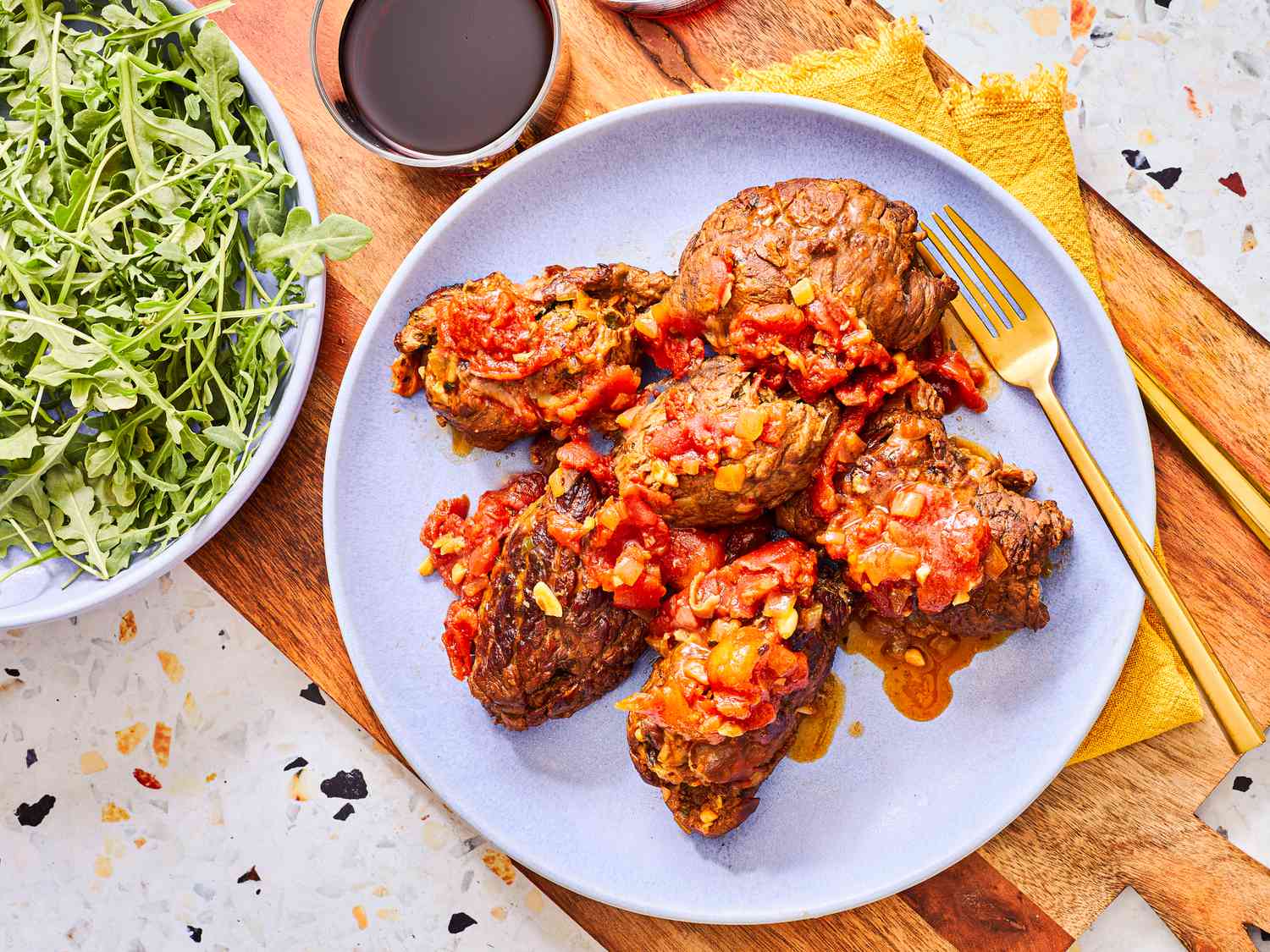 A platter of sliced Italian braciole arranged in a pool of rich tomato sauce, garnished with parsley on a rustic wooden table