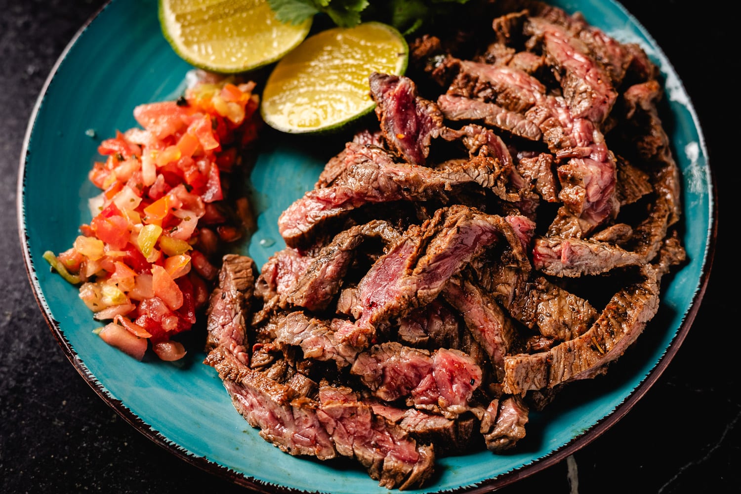 A platter of sliced carne asada with charred edges and lime wedges on a warm-toned wooden table, soft natural kitchen lighting