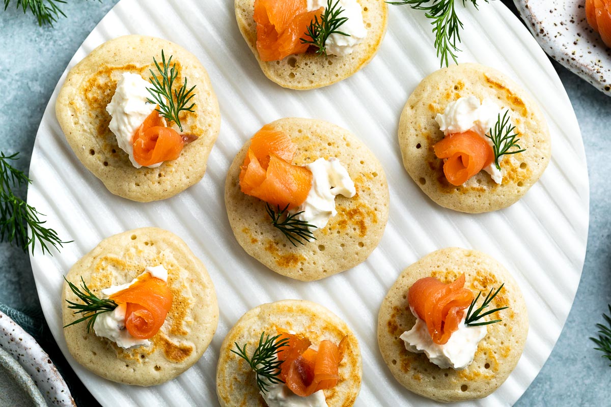 A platter of small golden blini topped with a curl of smoked salmon, a dollop of sour cream, and fresh dill on a linen-lined table setting