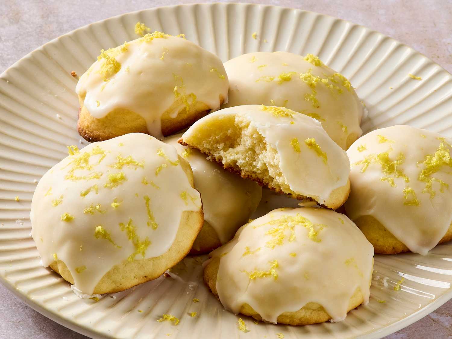 A platter of soft ricotta cookies topped with shiny lemon glaze on a light kitchen table with a small bowl of lemon zest nearby
