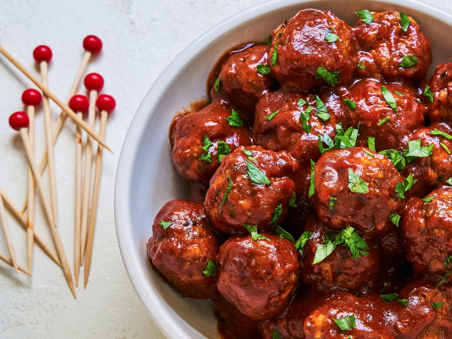 A platter of sticky BBQ-glazed meatballs with toothpicks on a warm wooden table, with a small bowl of extra sauce nearby and soft kitchen lighting